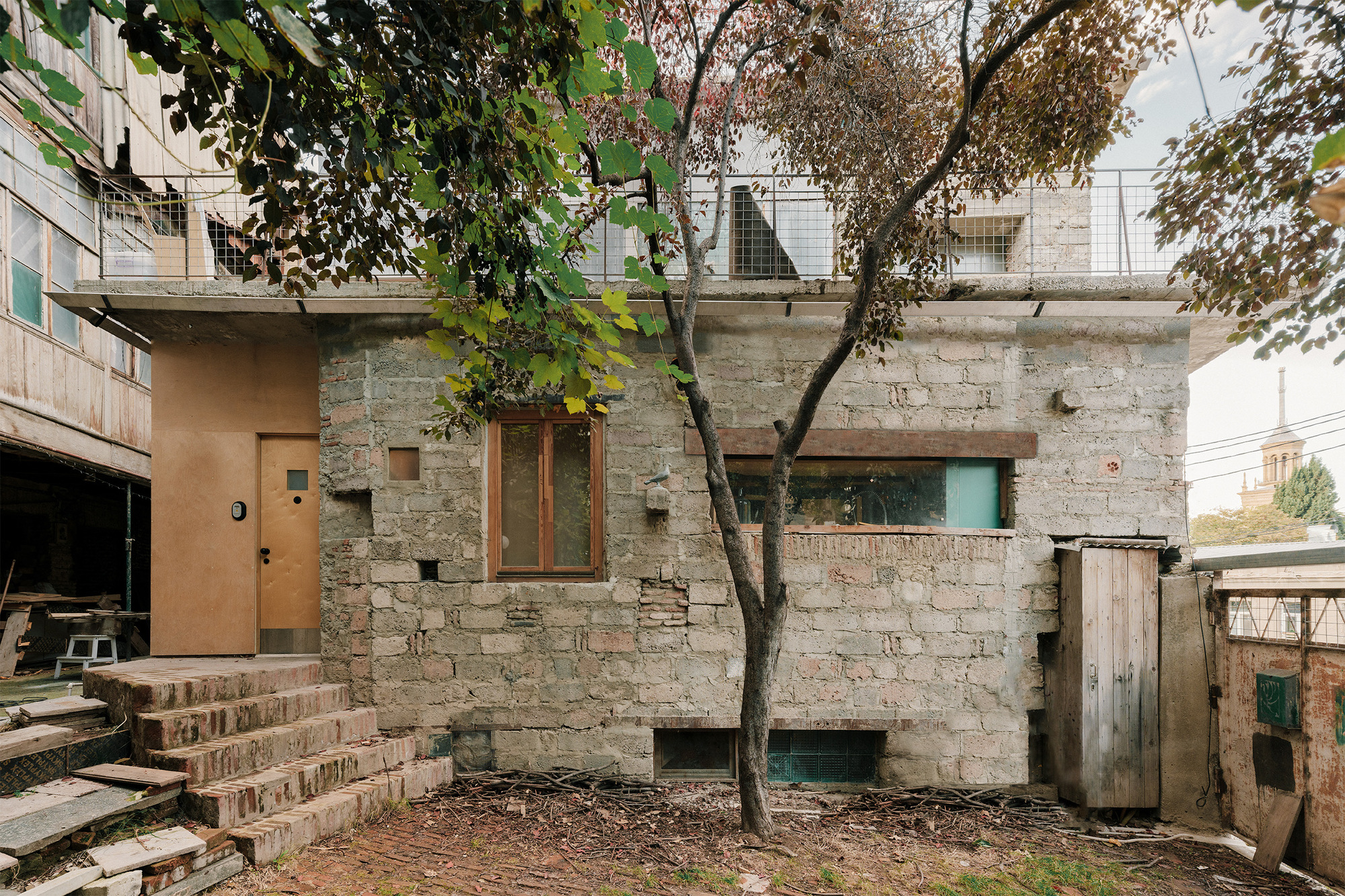 Stone block facade with timber window openings and a young tree in the courtyard below