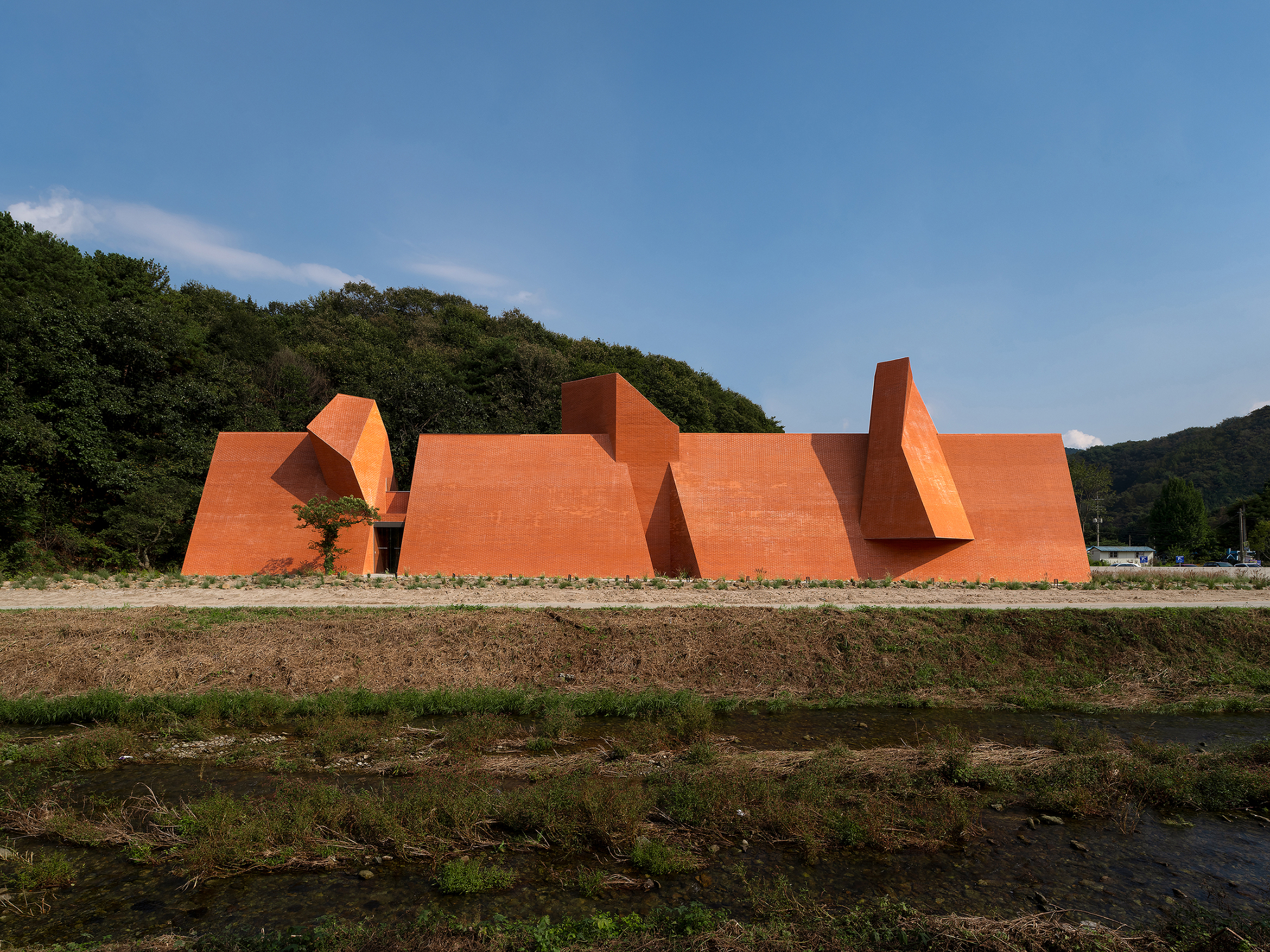 Long view of the angular orange brick volumes beside a forested hillside and plowed field