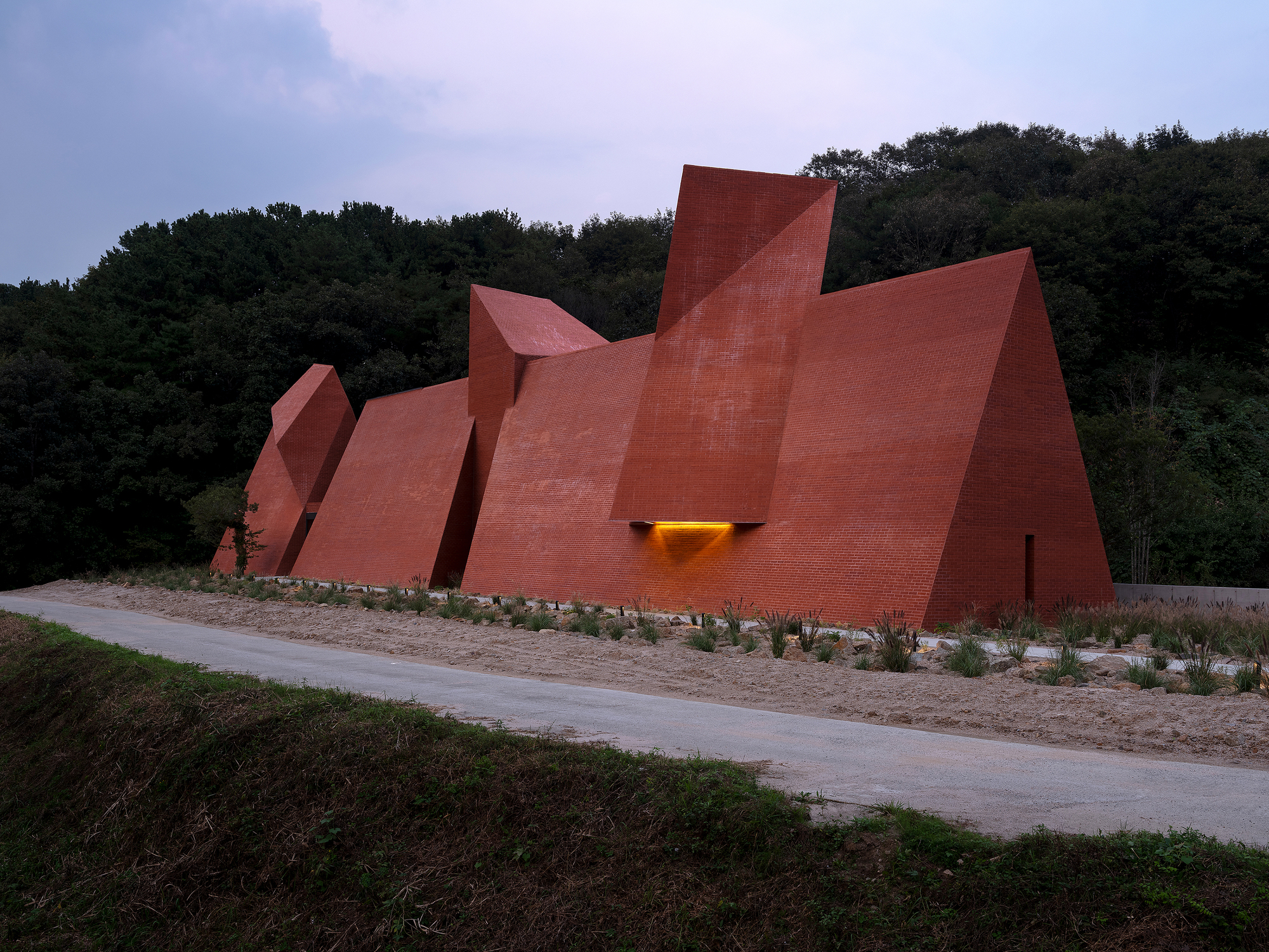 Angular brick facades at dusk with a single illuminated entrance opening beside the roadway