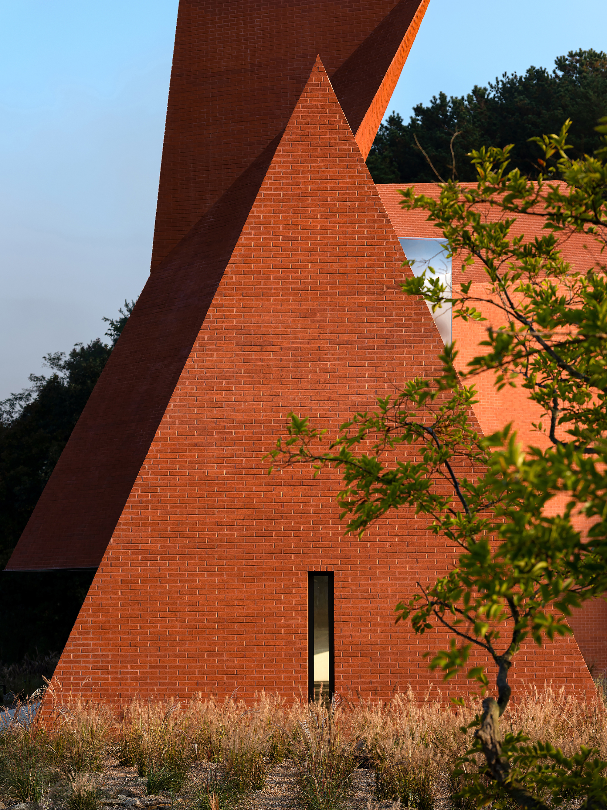 Triangular brick wall with a vertical slot window among autumn grasses and a young tree