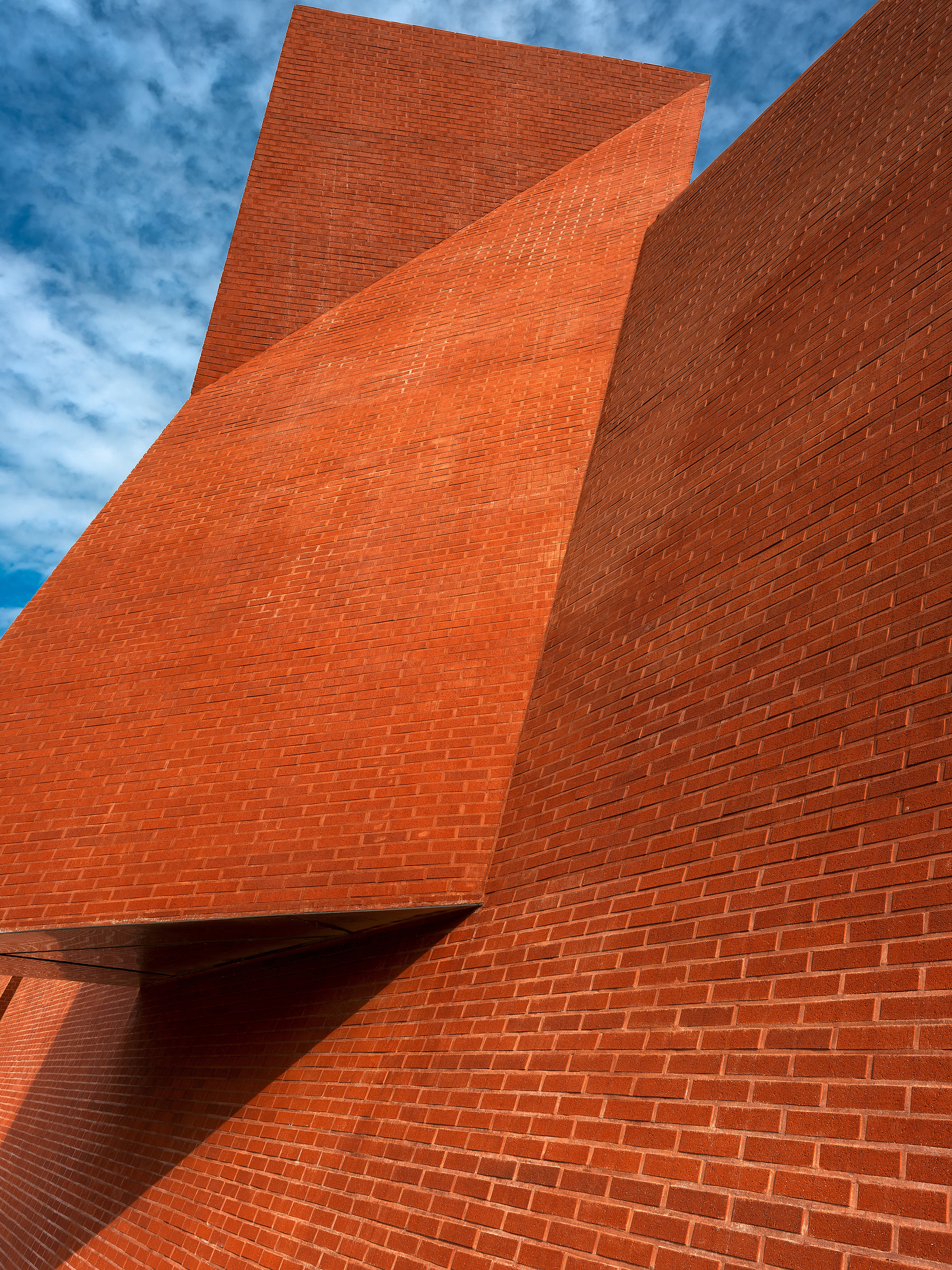 Upward view of folded red brick facade planes meeting at sharp angles under blue sky