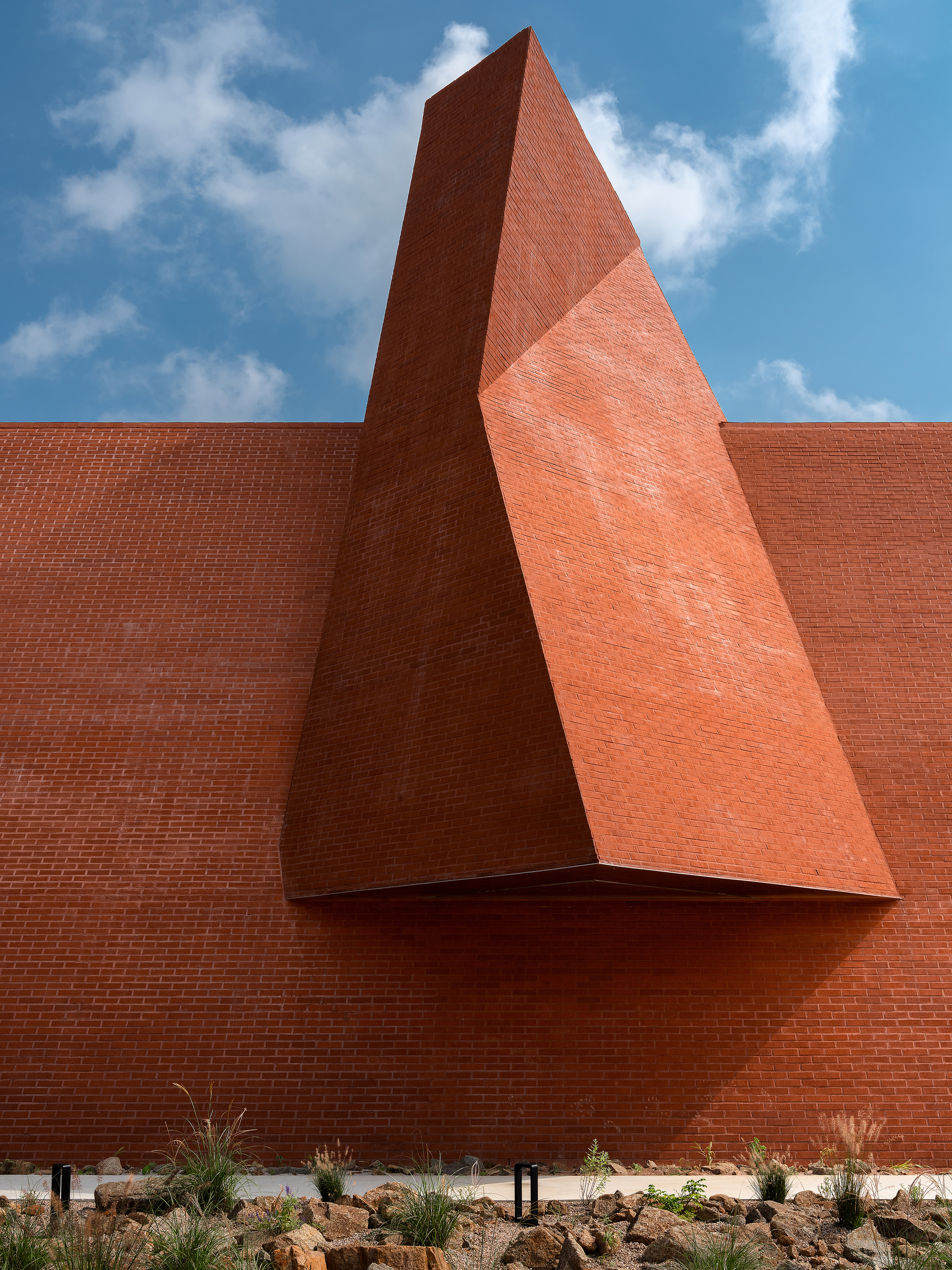 Triangular red brick volume projecting from flat wall above native grasses and rock garden