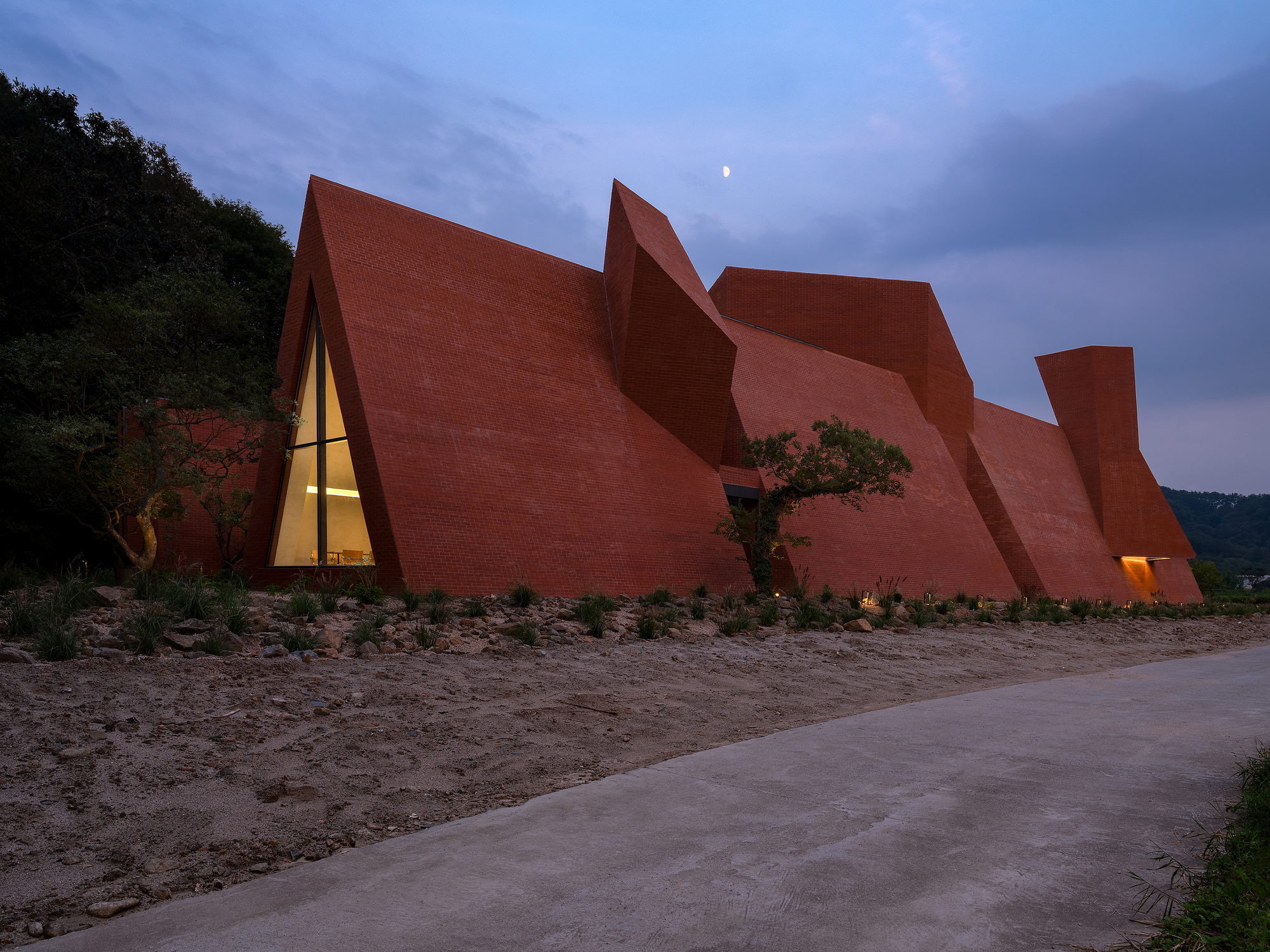 Angular red brick facade with triangular glass openings glowing at twilight beside a concrete path