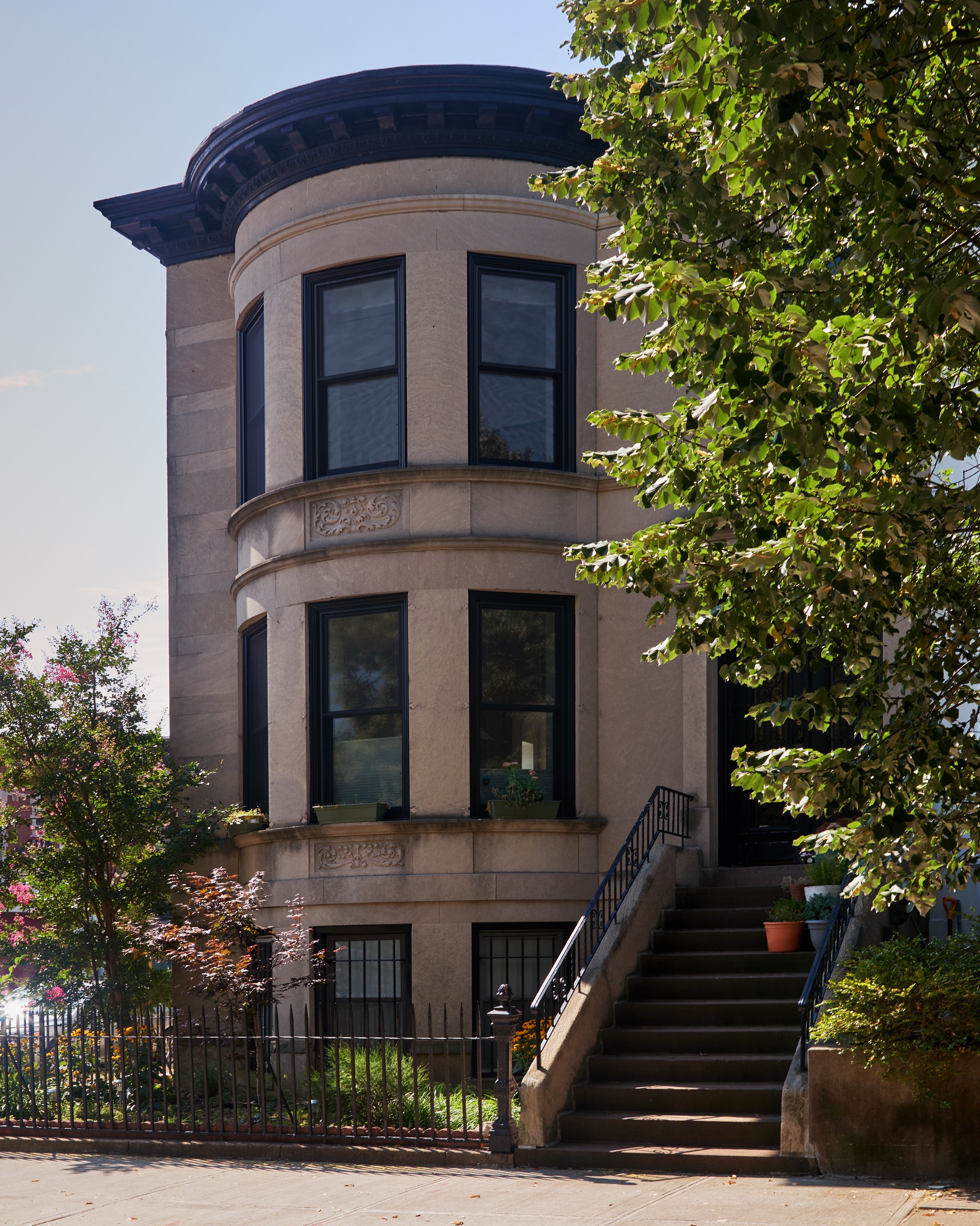 Curved facade of sandstone rowhouse with bay windows and entry stair flanked by trees