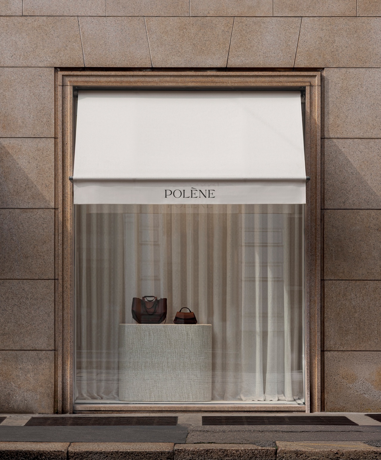 Single window display framed in bronze with white awning and sheer curtains against stone panels