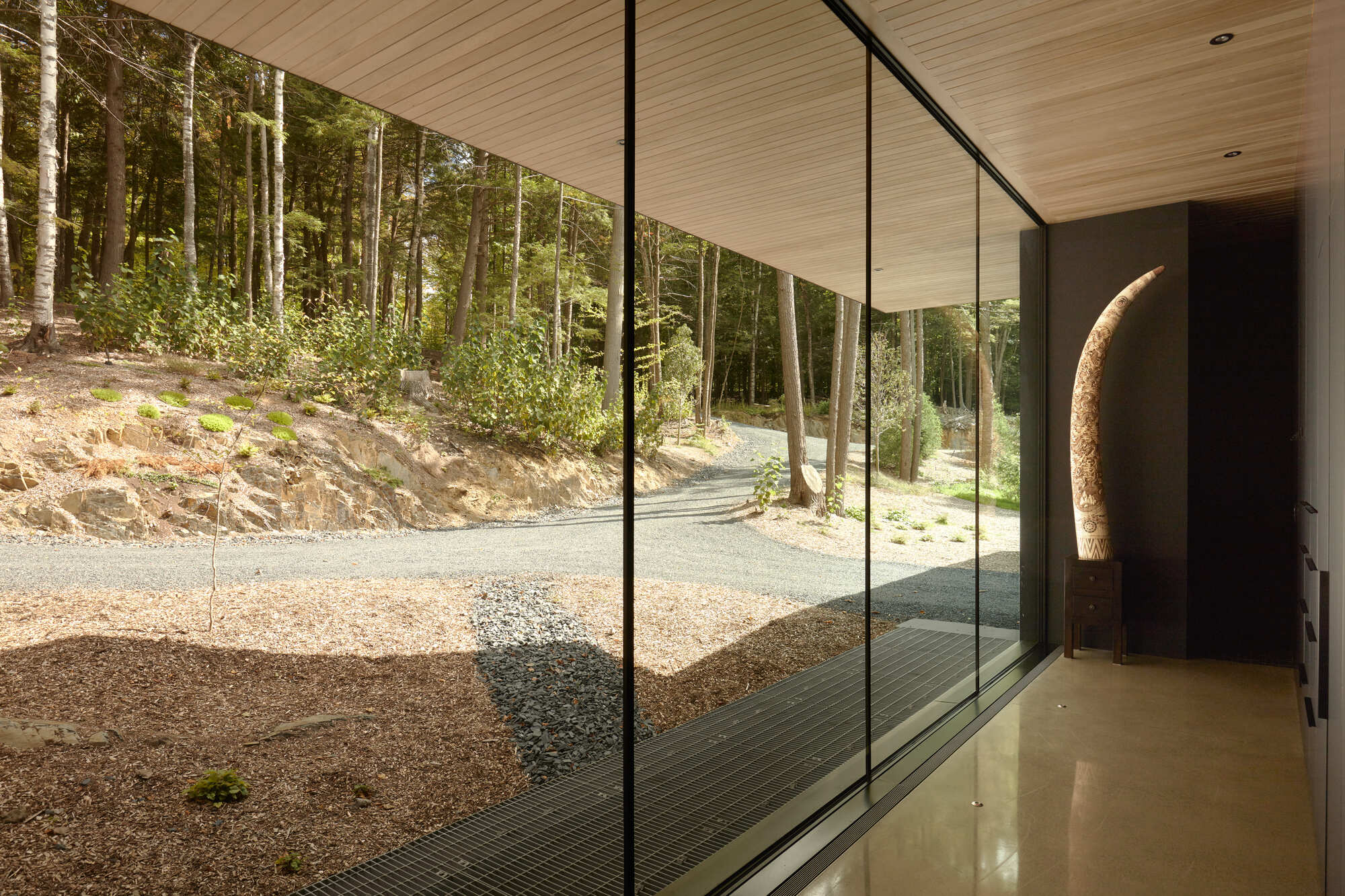 Glass-walled corridor with timber ceiling overlooking a gravel courtyard and forested hillside