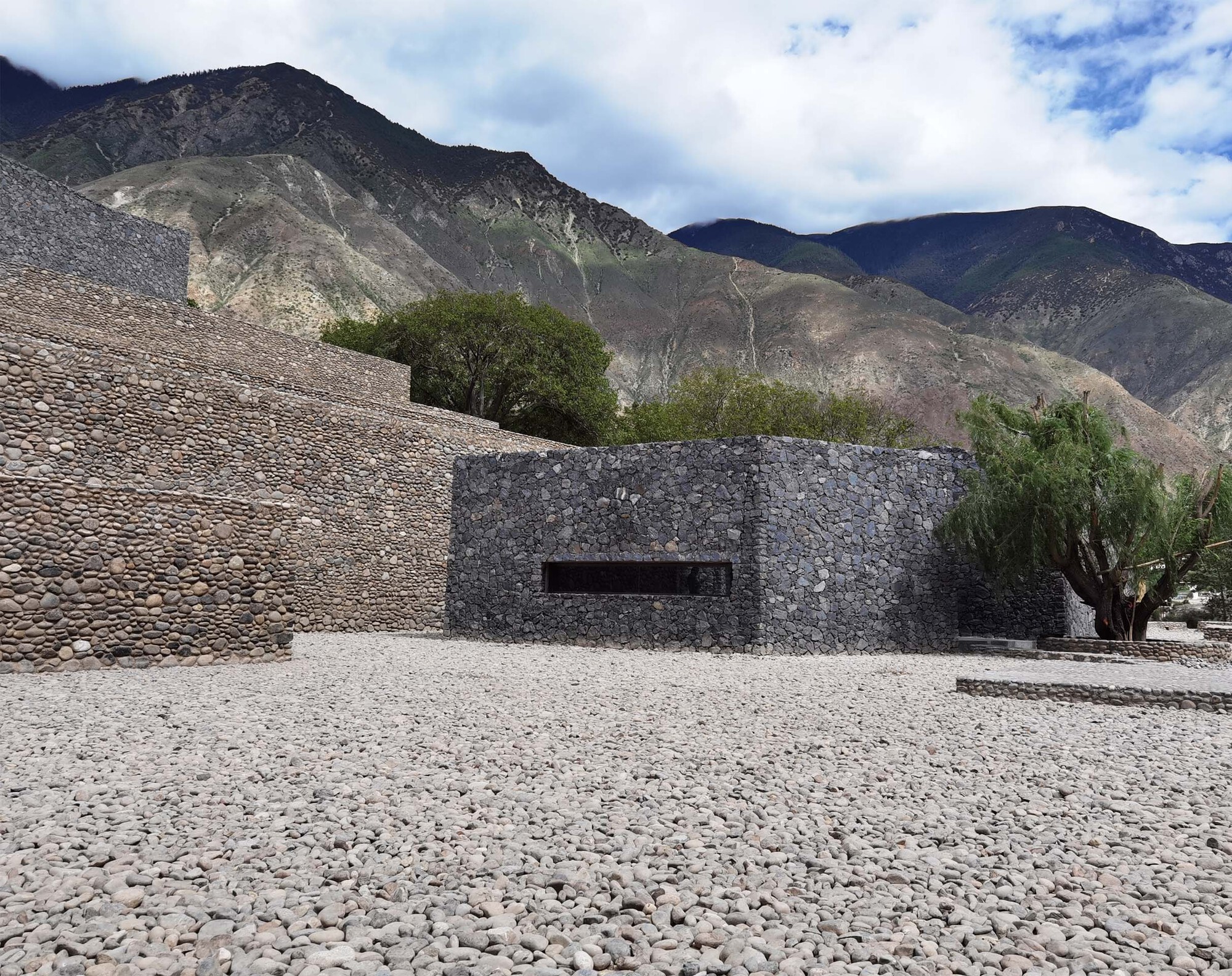 Stone gabion facade with horizontal window opening against a pebbled plaza and mountain backdrop