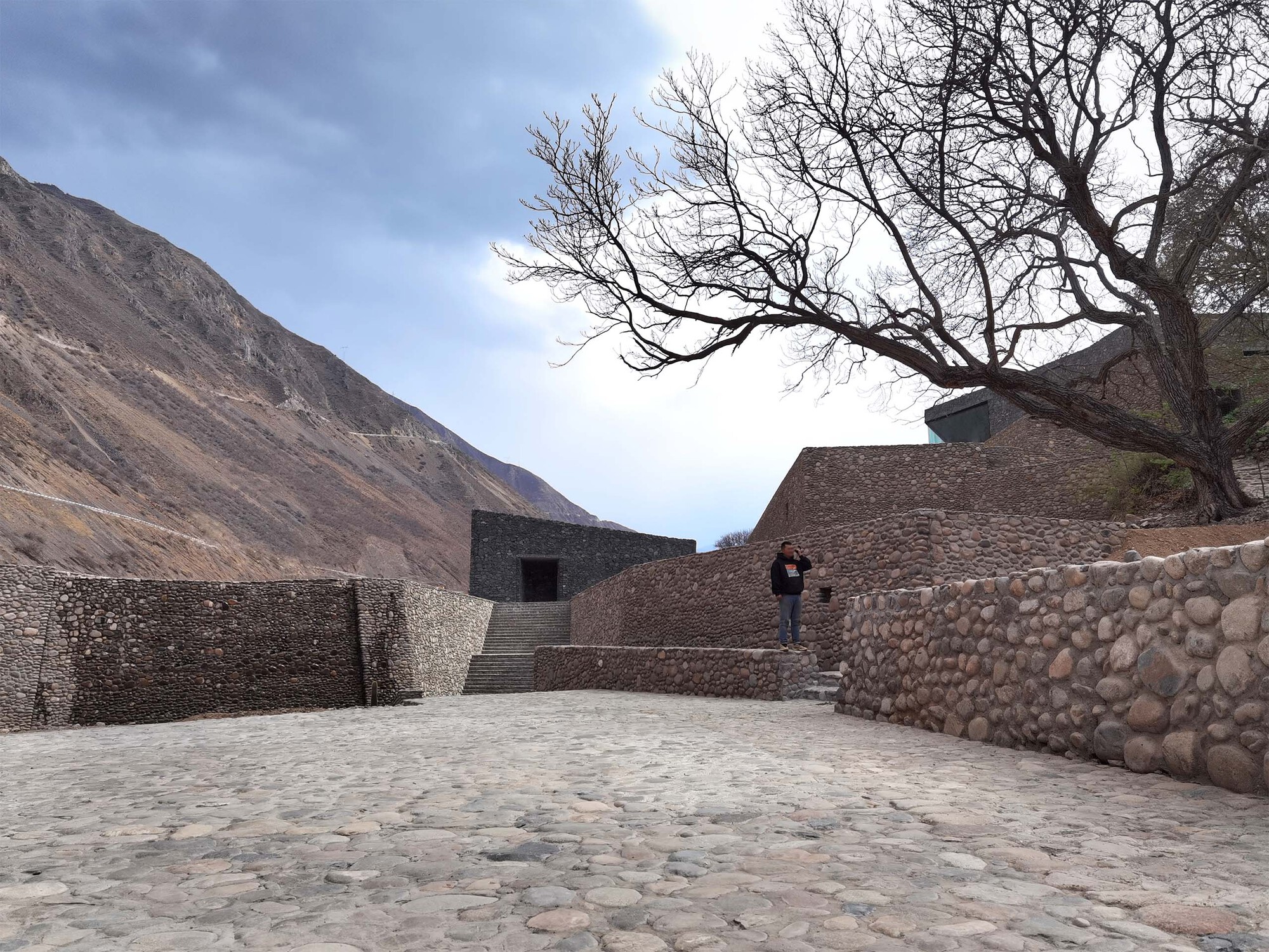 Cobbled courtyard enclosed by stone gabion walls with a figure ascending stairs under a leafless tree