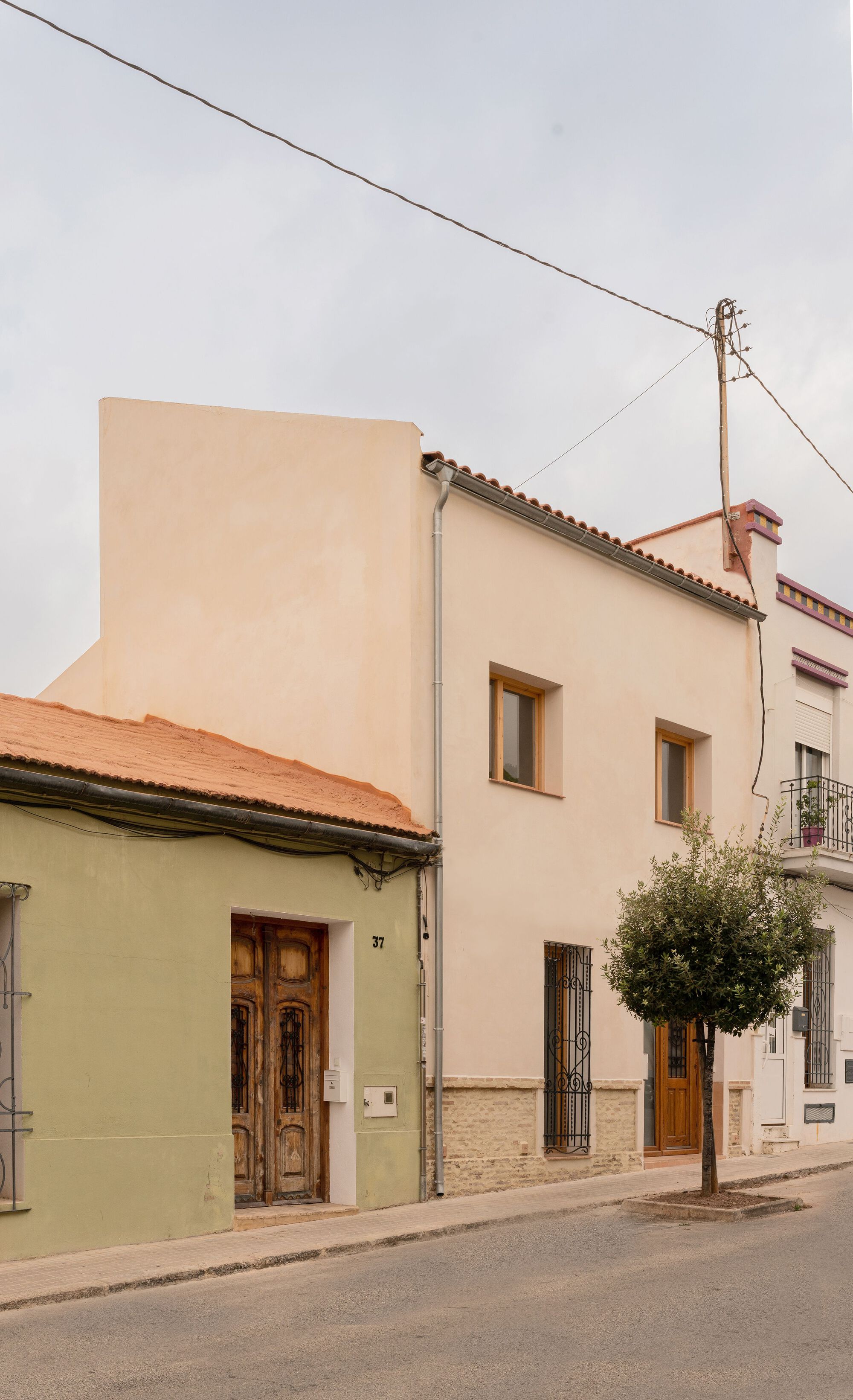Street facade showing beige plaster volume with barrel tile roof beside a small street tree