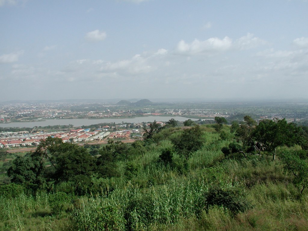 Elevated view from a vegetated hillside overlooking low-rise residential developments and a river in hazy daylight