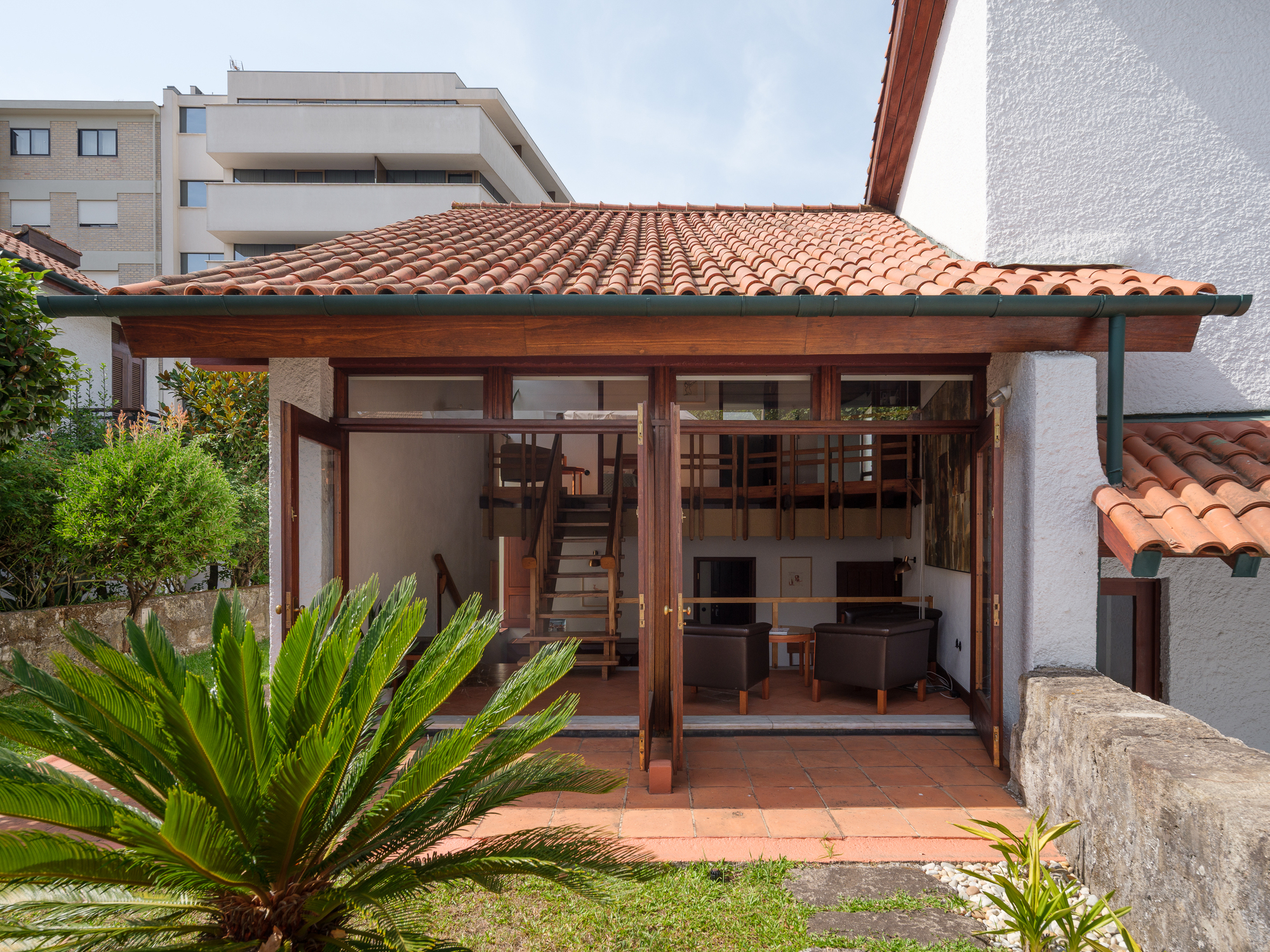 Garden-facing pavilion with timber-framed glass doors and terracotta tile roof framed by palm fronds