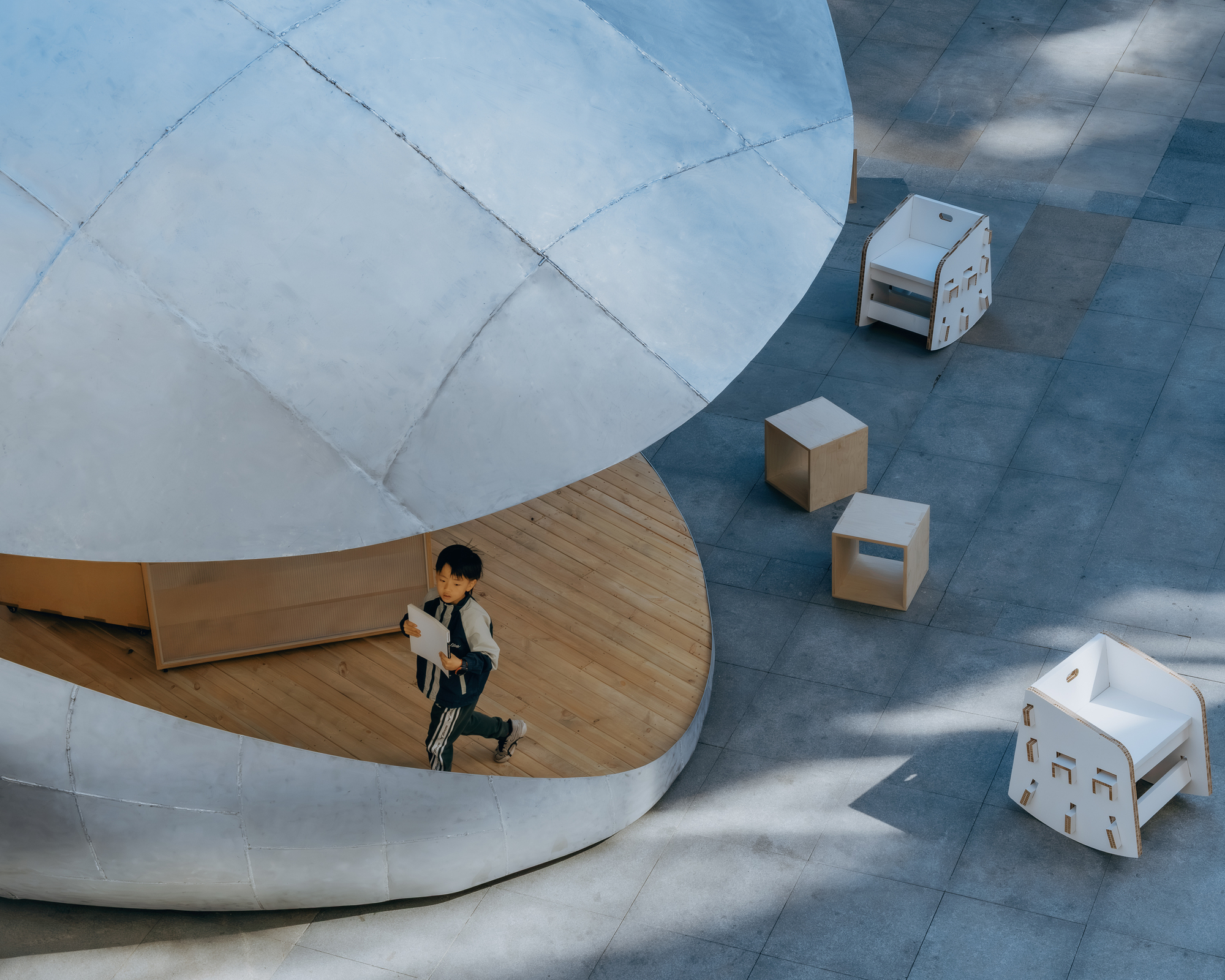 Overhead view of a child on the curved plywood bench with white cardboard stools on the paved plaza