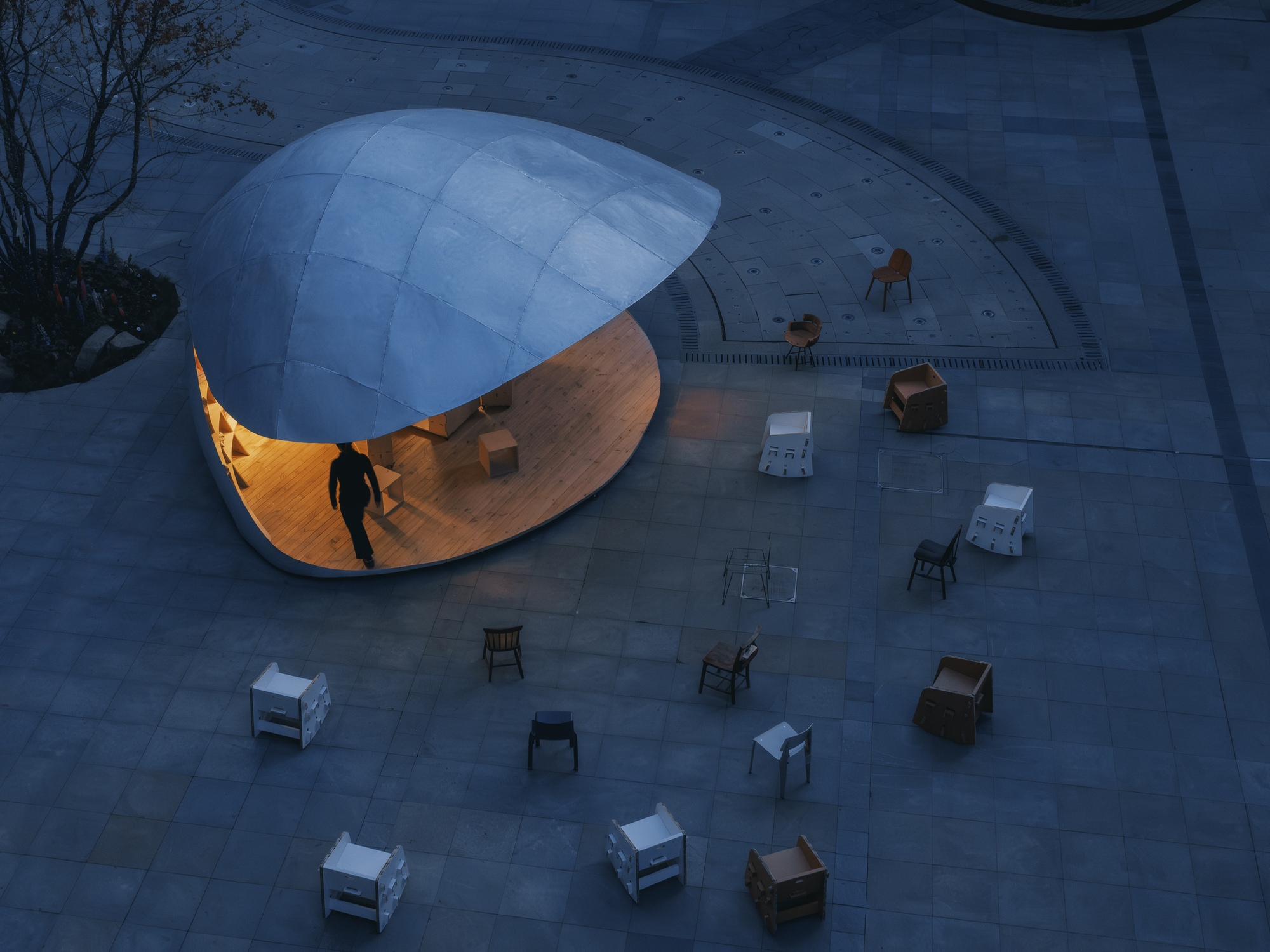 Aerial view of the illuminated pavilion at dusk surrounded by scattered furniture on paved plaza