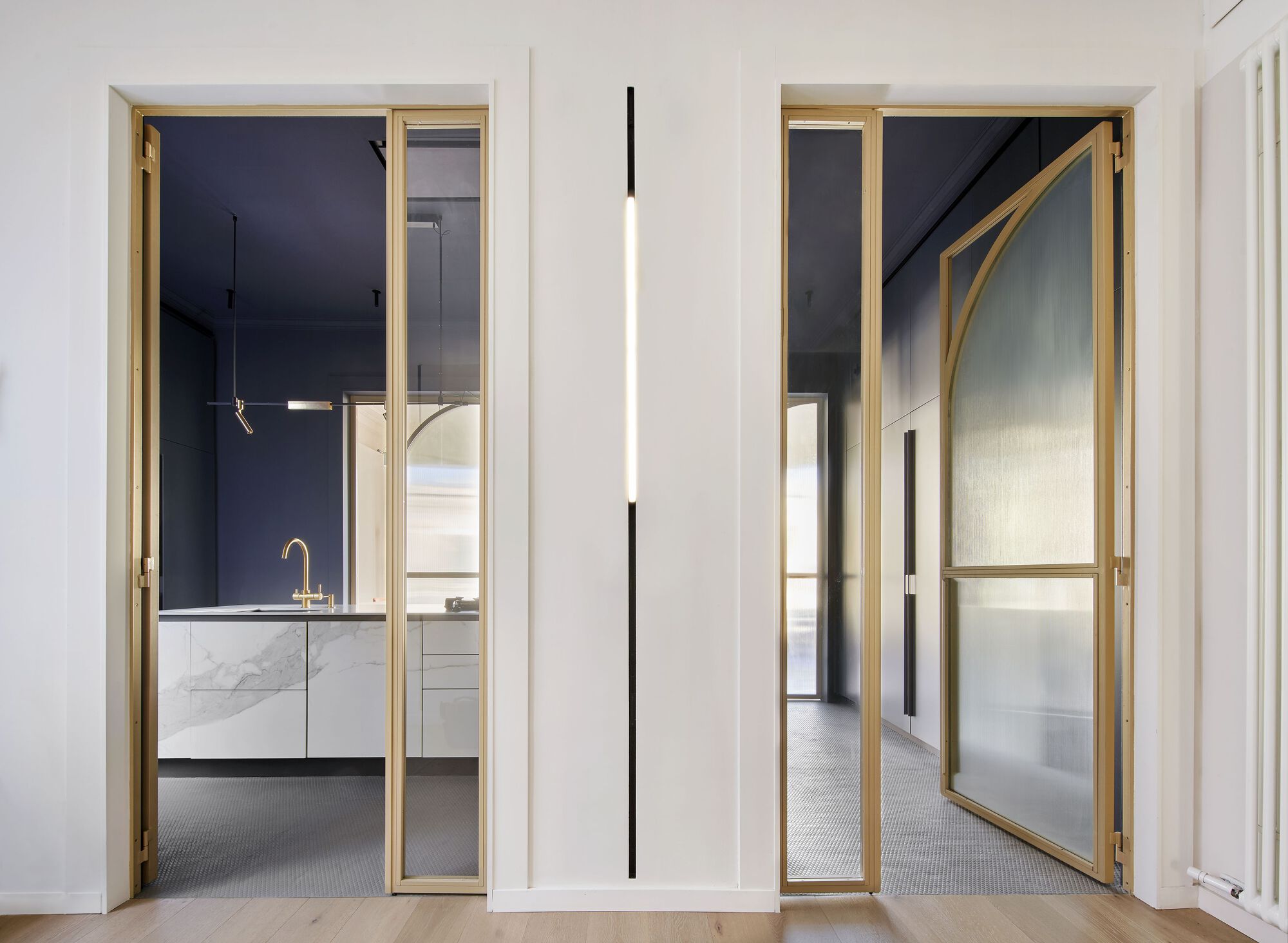 Brass-framed pivoting door with frosted glass panel and arched structural brace leading to kitchen beyond