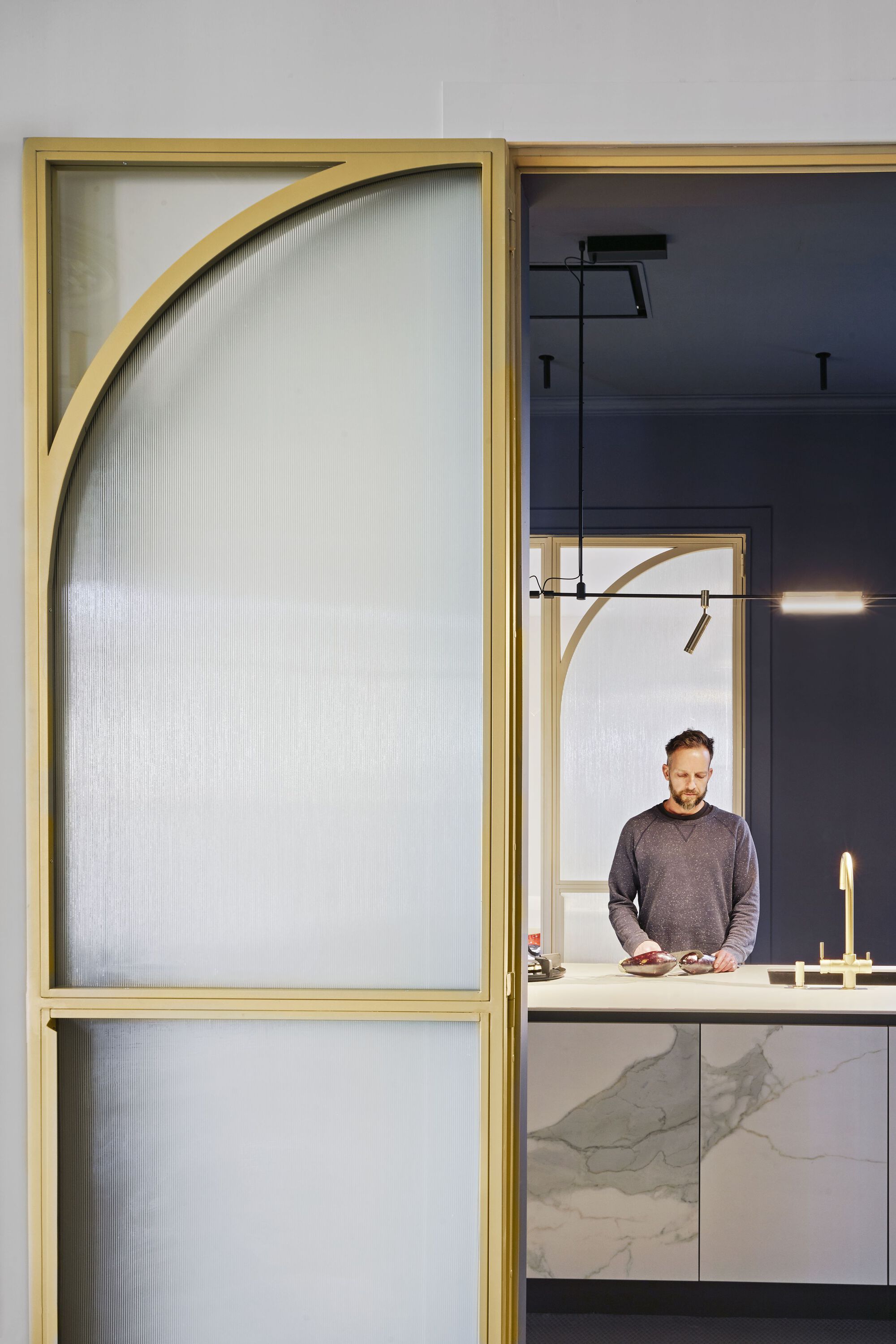 Close-up of brass-framed translucent door panel with curved brace and resident working at marble kitchen island