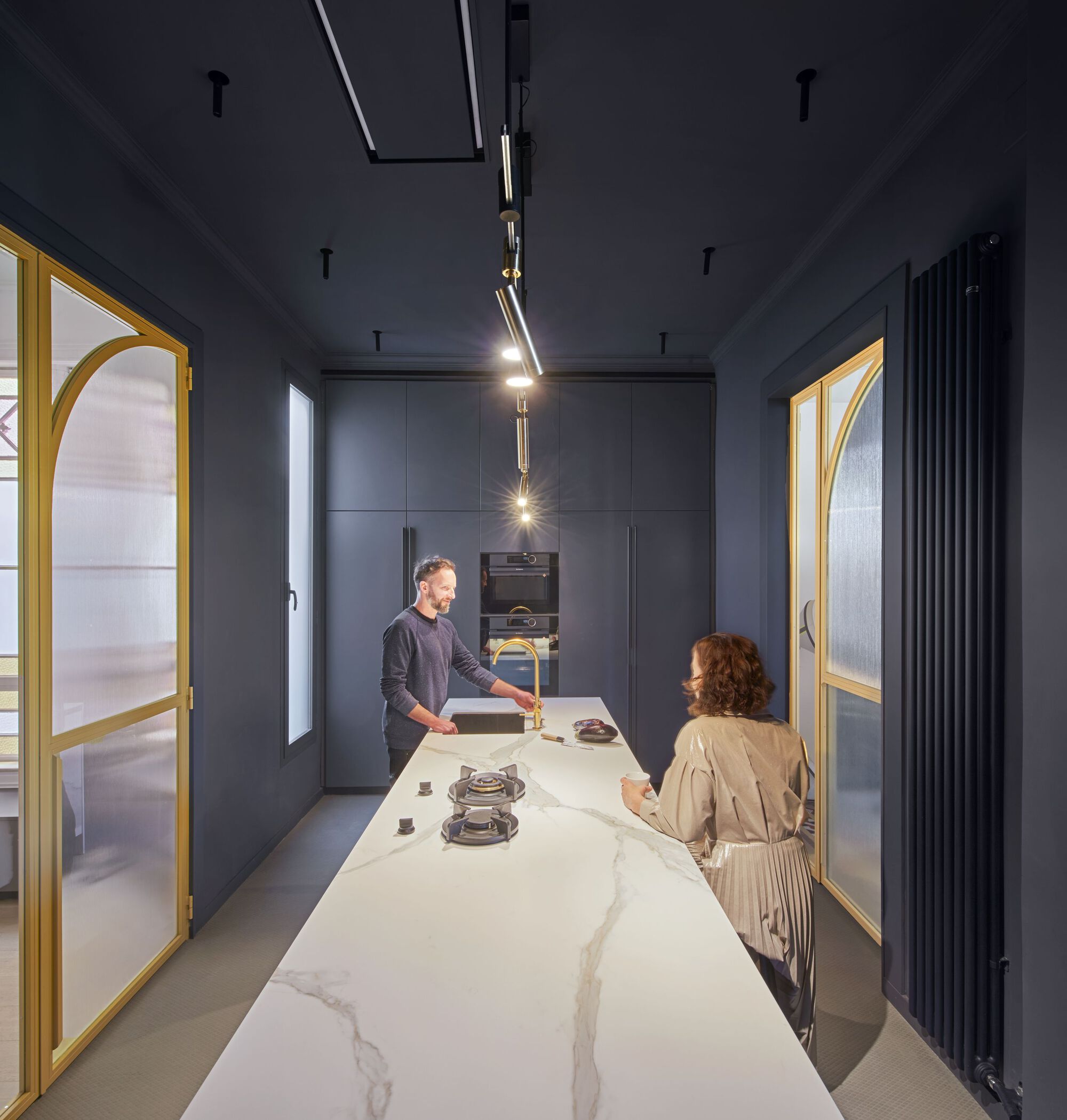 Galley kitchen with dark blue cabinetry, marble island, and two residents under articulated linear pendant light