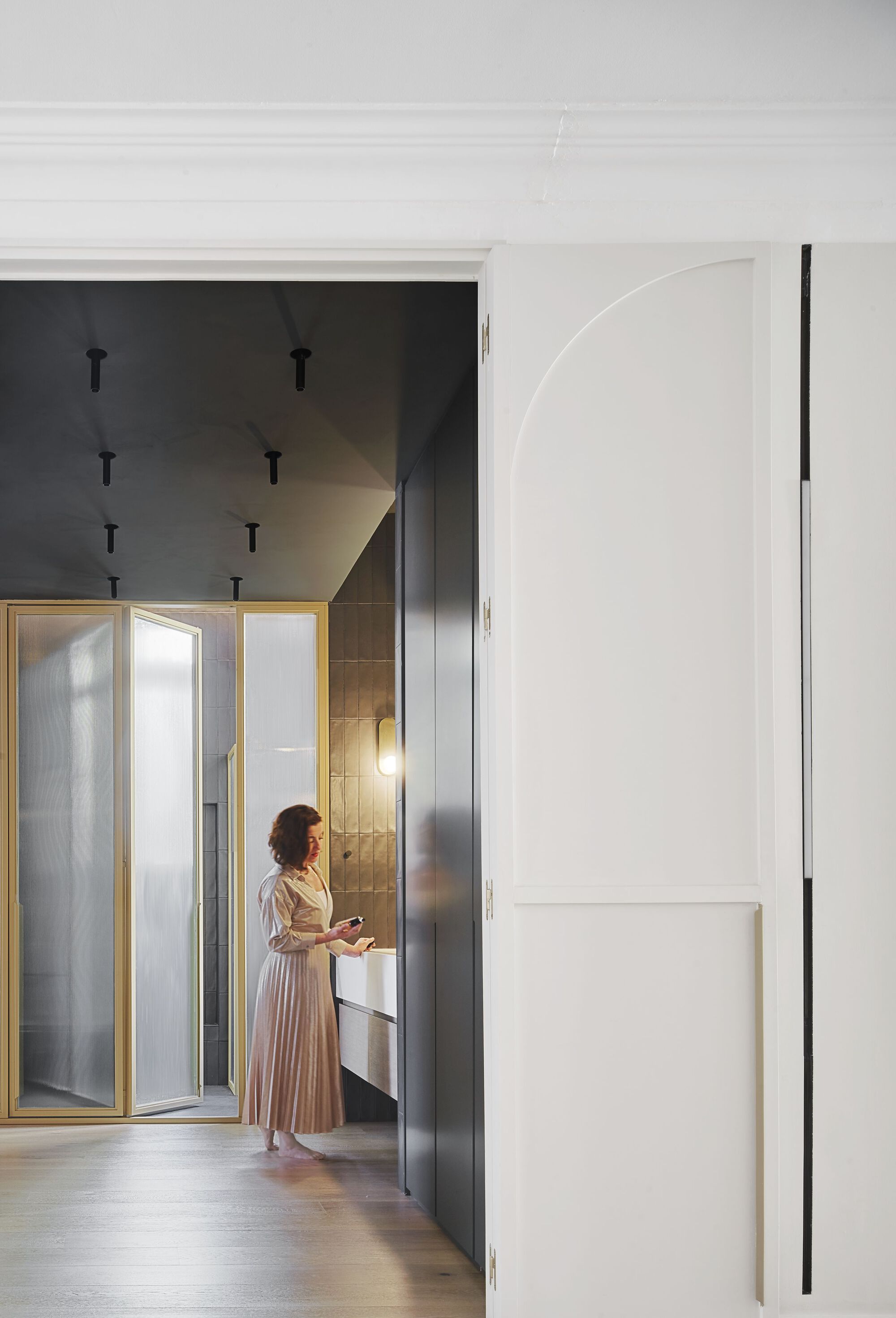 Woman in a pleated dress standing beside dark grey cabinetry and brass-framed translucent glass doors