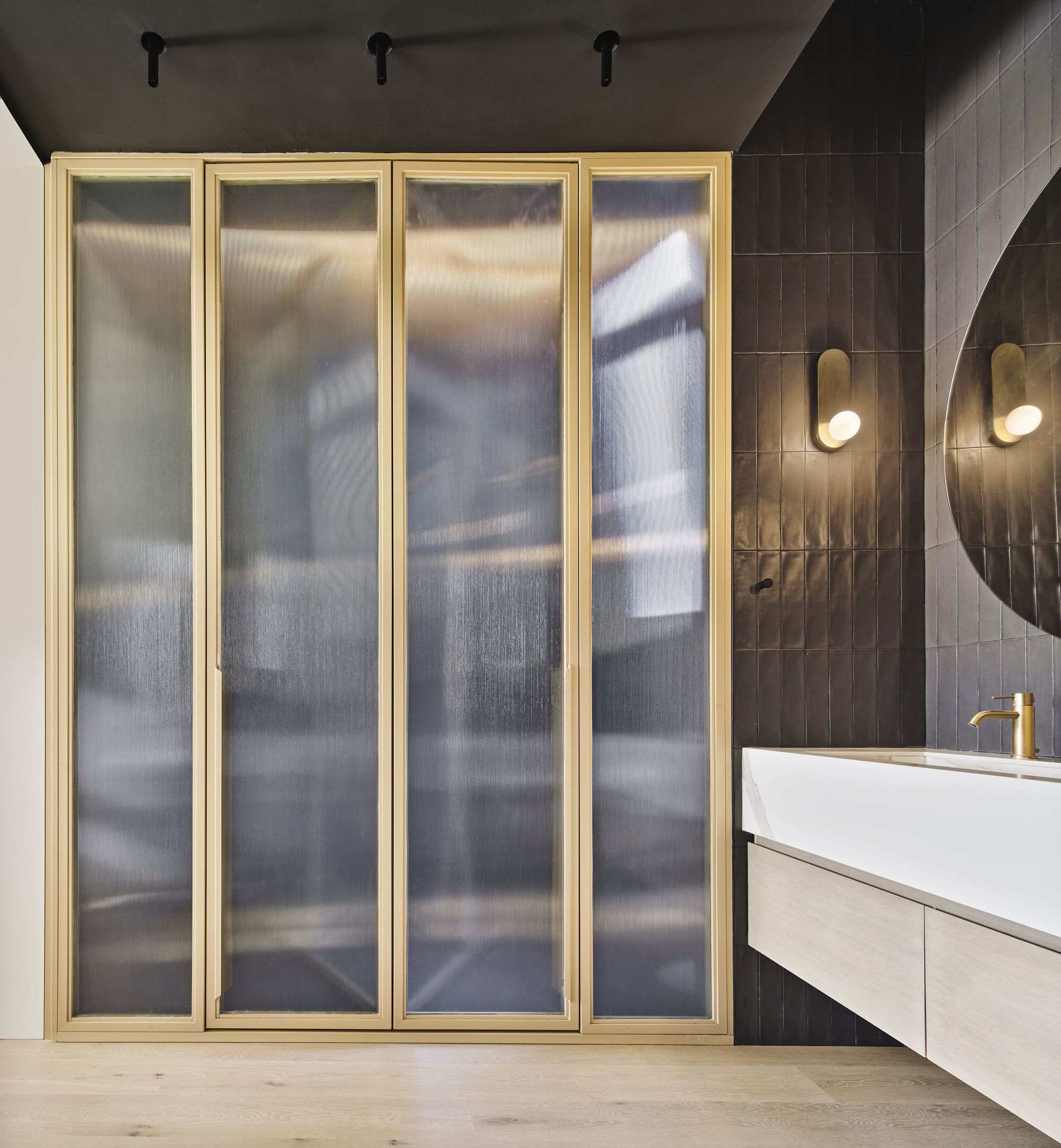 Brass-framed translucent glass folding doors adjacent to dark tiled bathroom wall with circular mirror and sconces