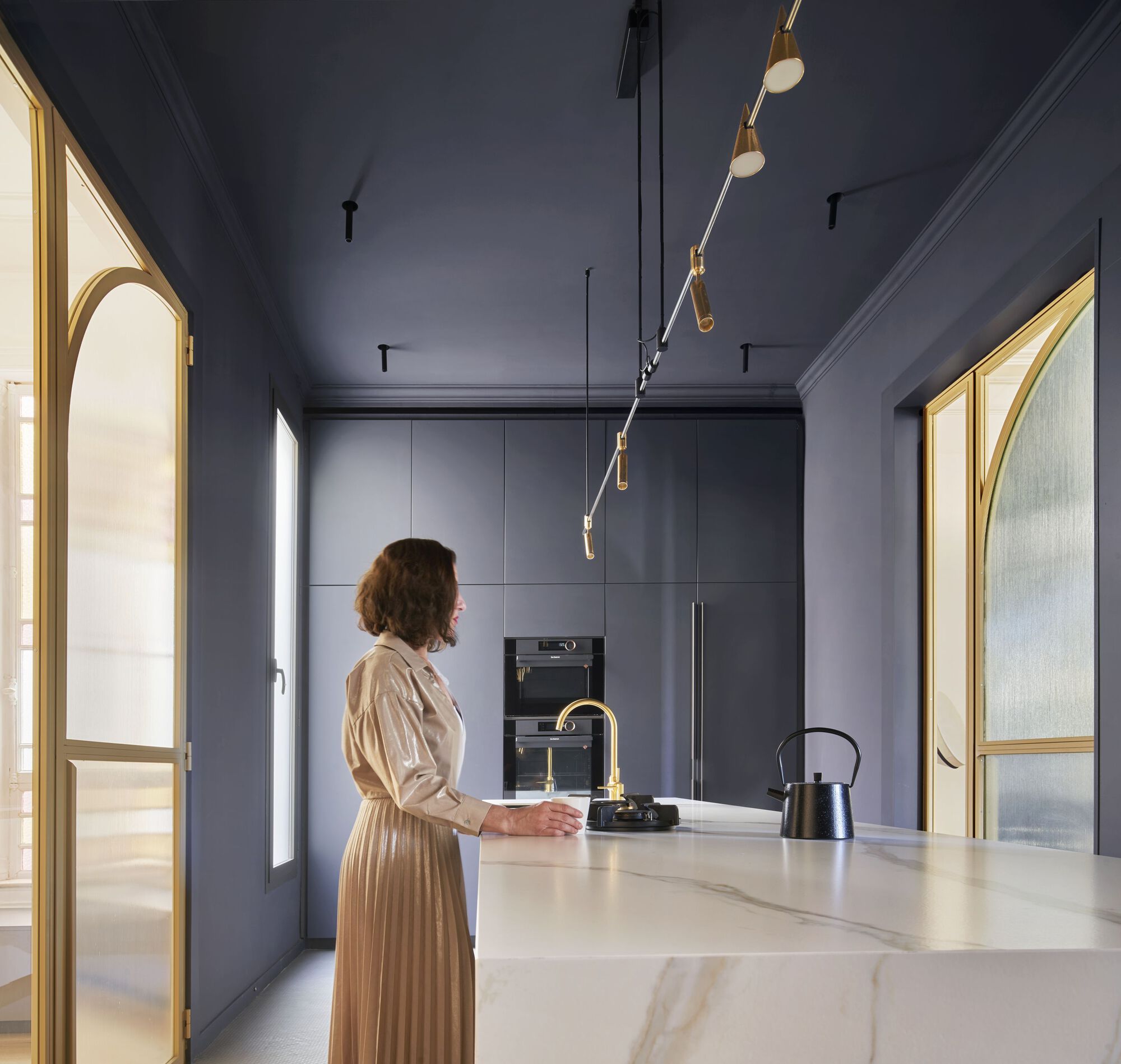 Kitchen with slate blue cabinetry and marble island where a person stands by the brass faucet