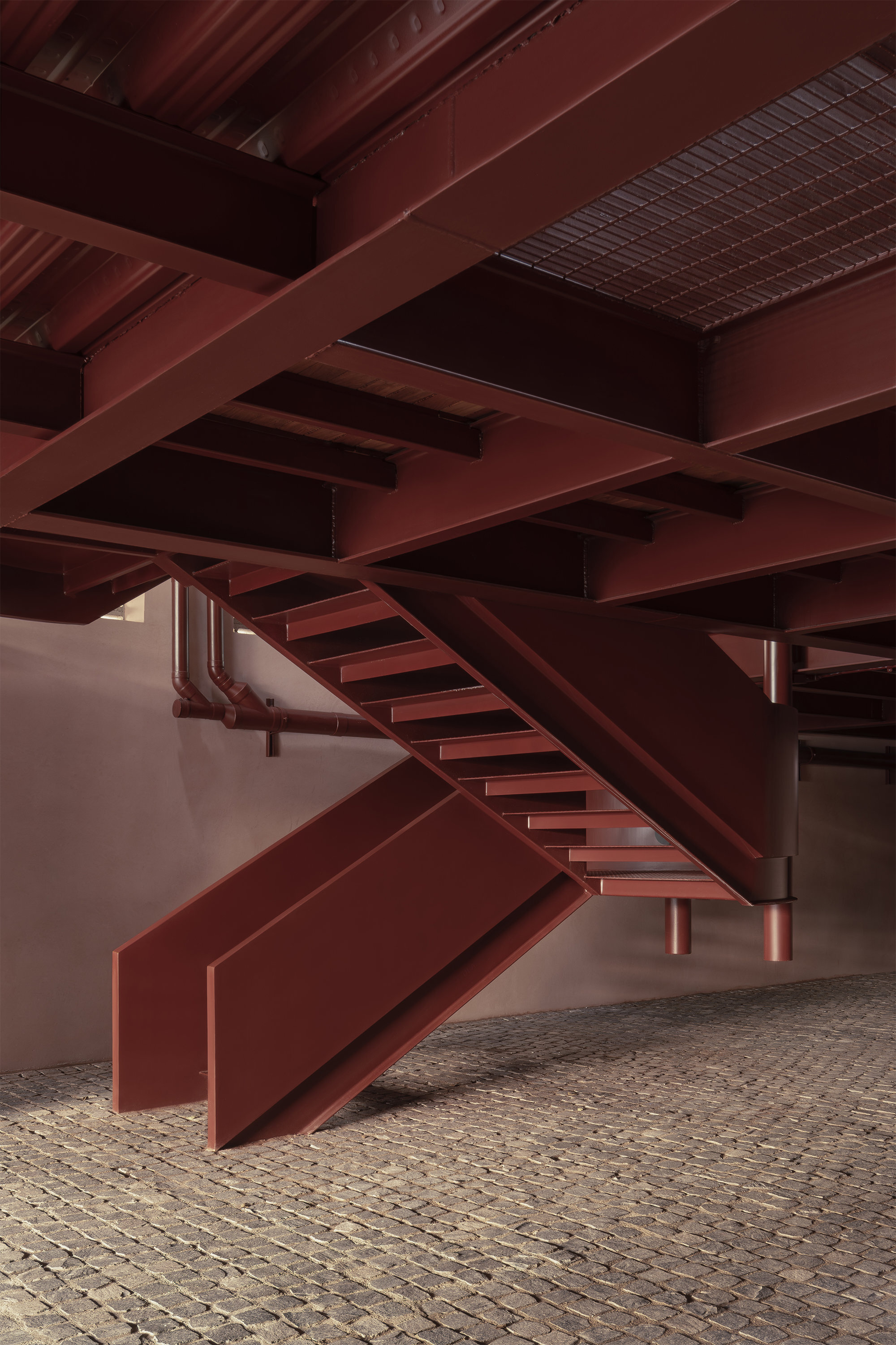 Underside view of intersecting steel beams and staircases with exposed structural grid above cobblestones