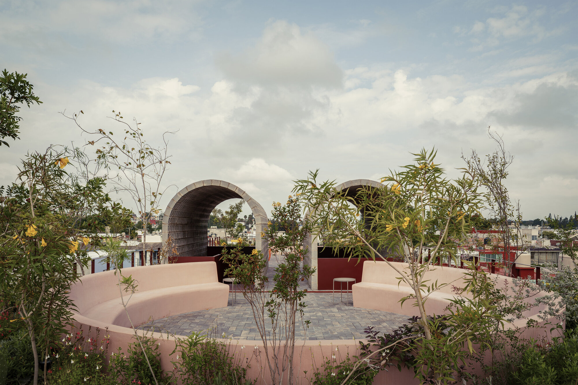 Rooftop terrace with pink curved seating walls and twin barrel vault arches framed by planted beds under cloudy skies