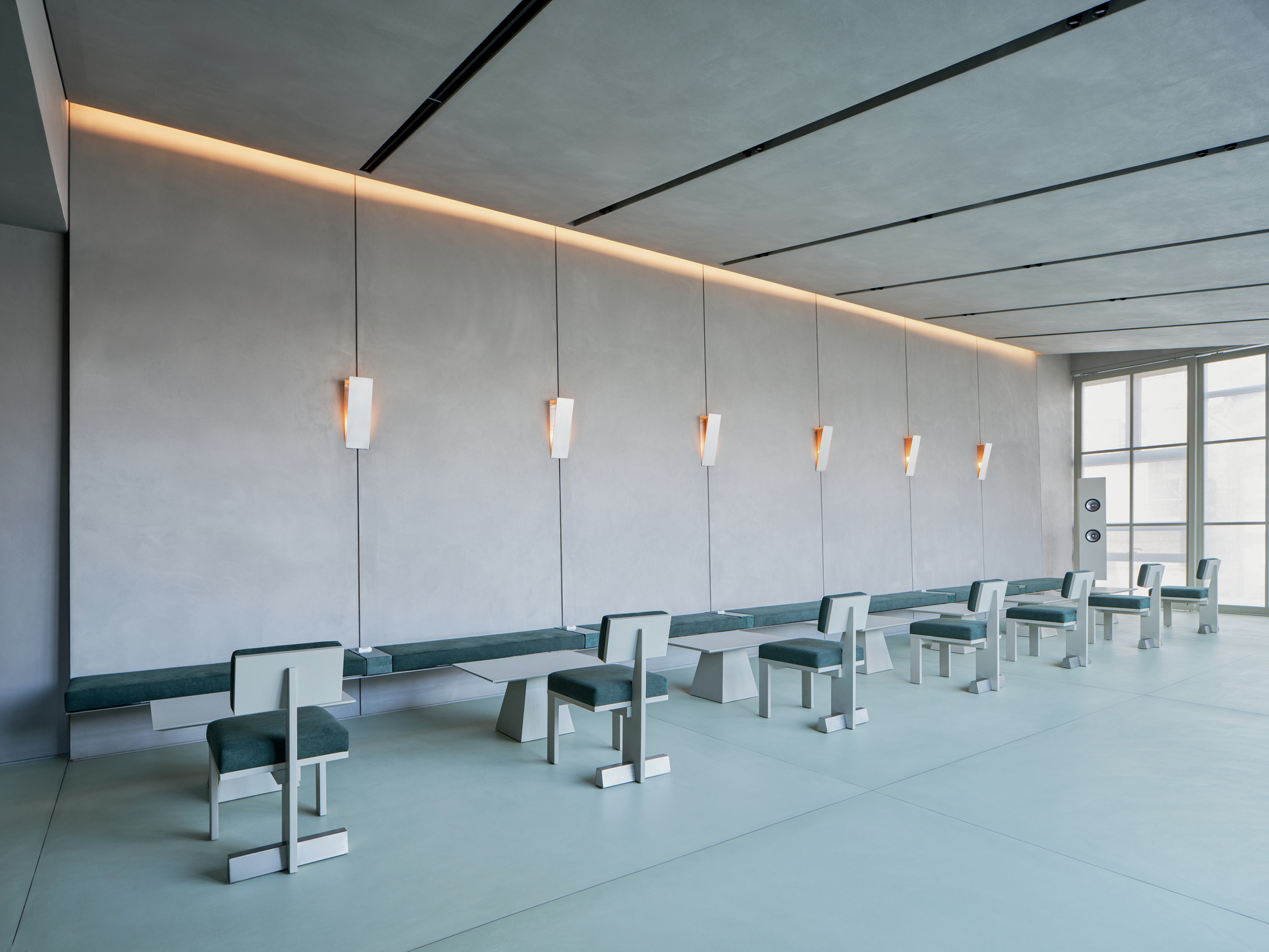 Interior meditation hall with rows of teal benches and suspended pendant lights along a grey paneled wall