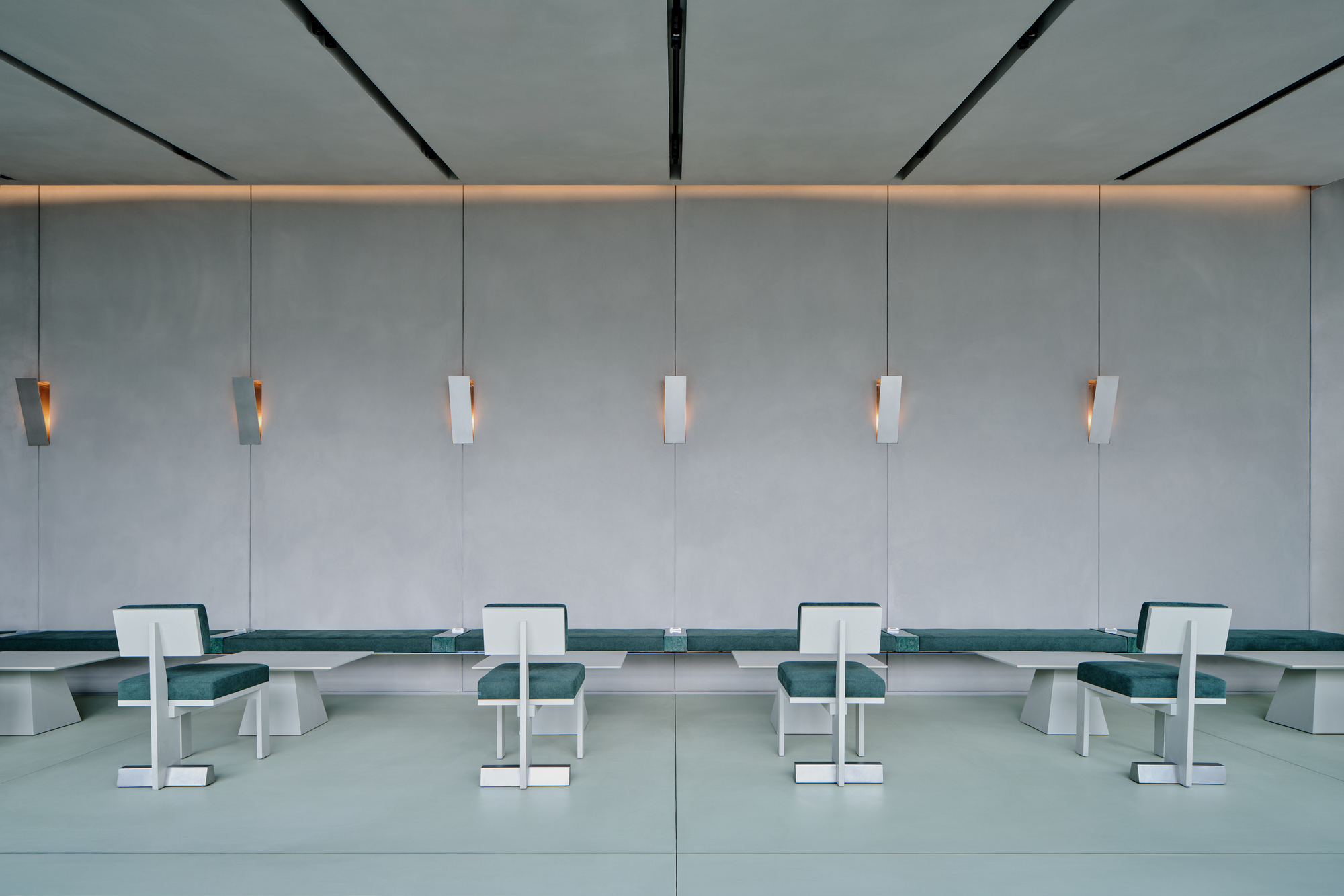 Symmetrical wall of grey panels with six suspended pendant lights above teal upholstered benches and white chairs