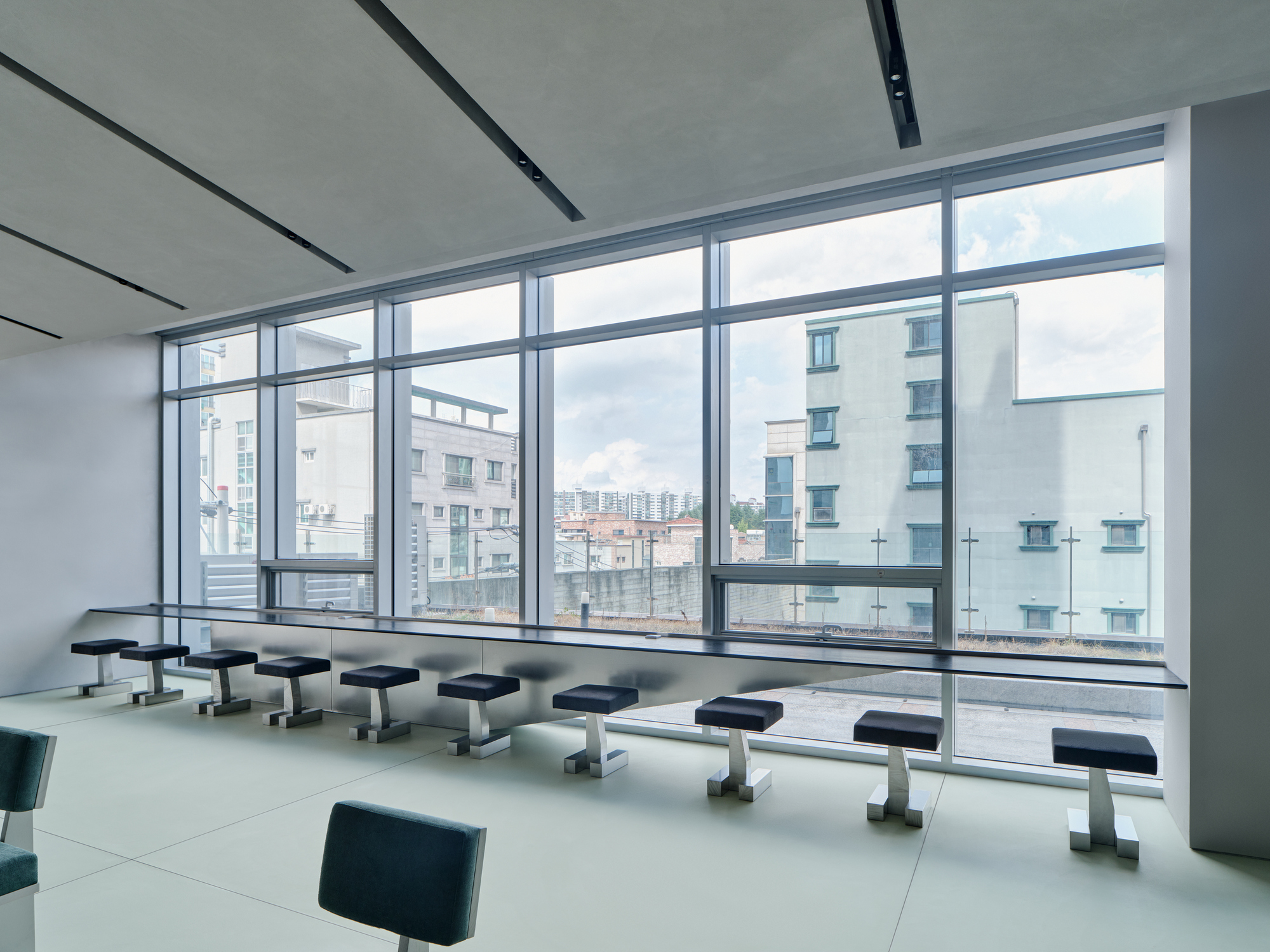 Row of dark upholstered stools facing floor-to-ceiling windows with views of surrounding residential buildings