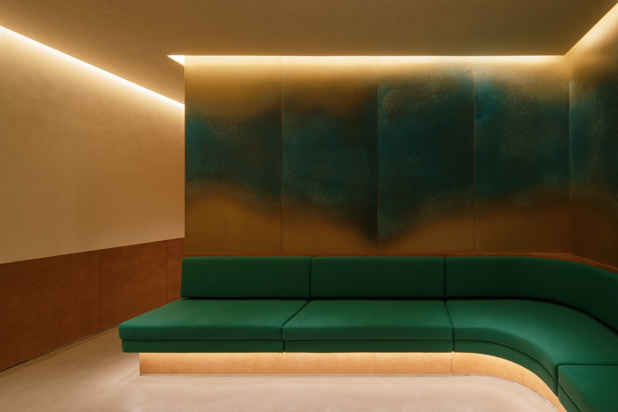Recessed cove lighting above a curved green velvet banquette against gradient painted wall panels