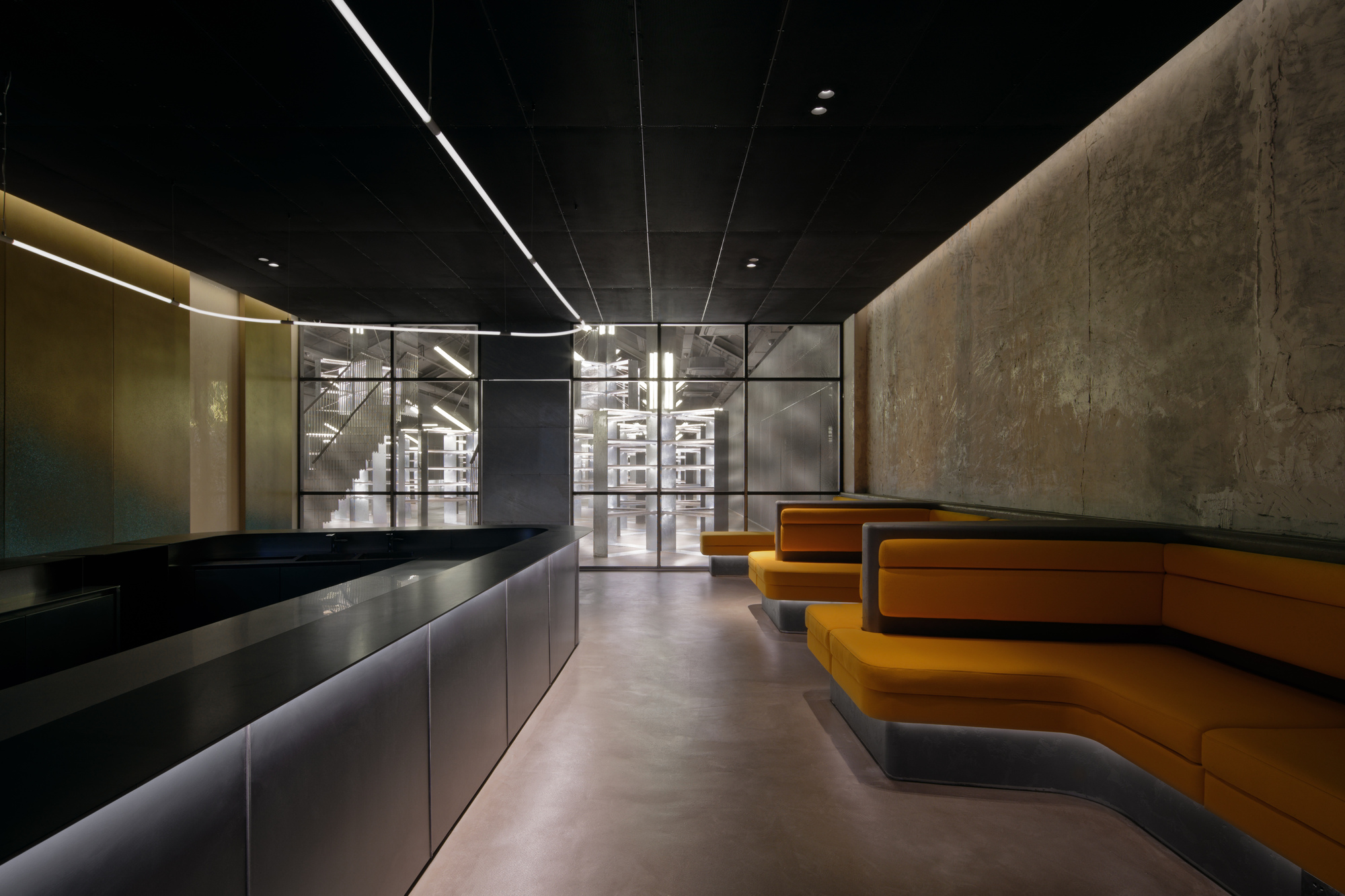 Reception lobby with black tiered counter, textured concrete wall and orange stepped seating under linear ceiling lights