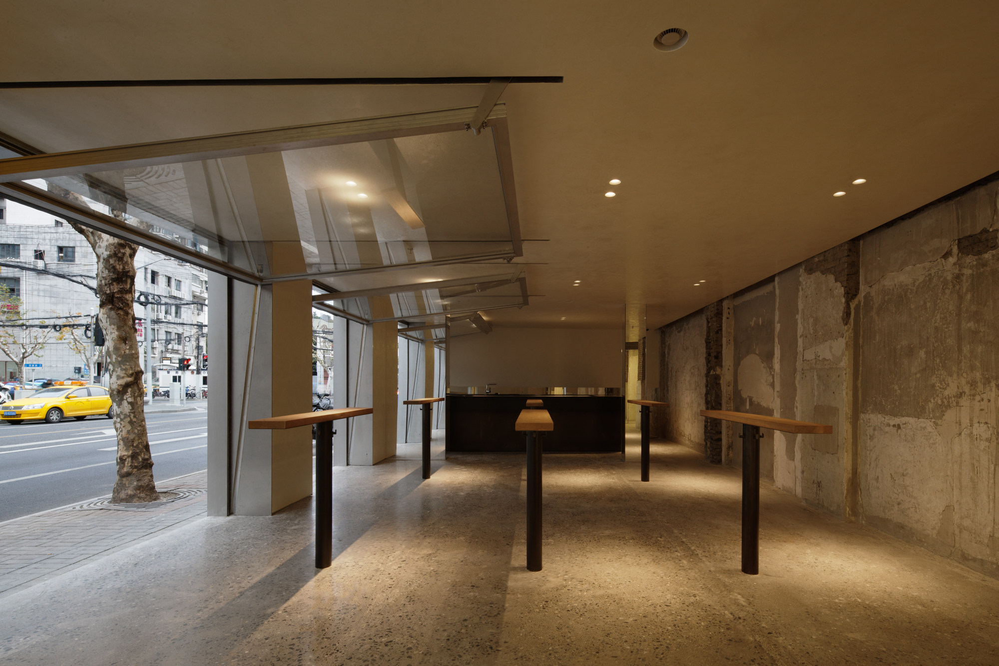 Ground floor interior with street trees visible through glass facade and standing height work tables on terrazzo flooring