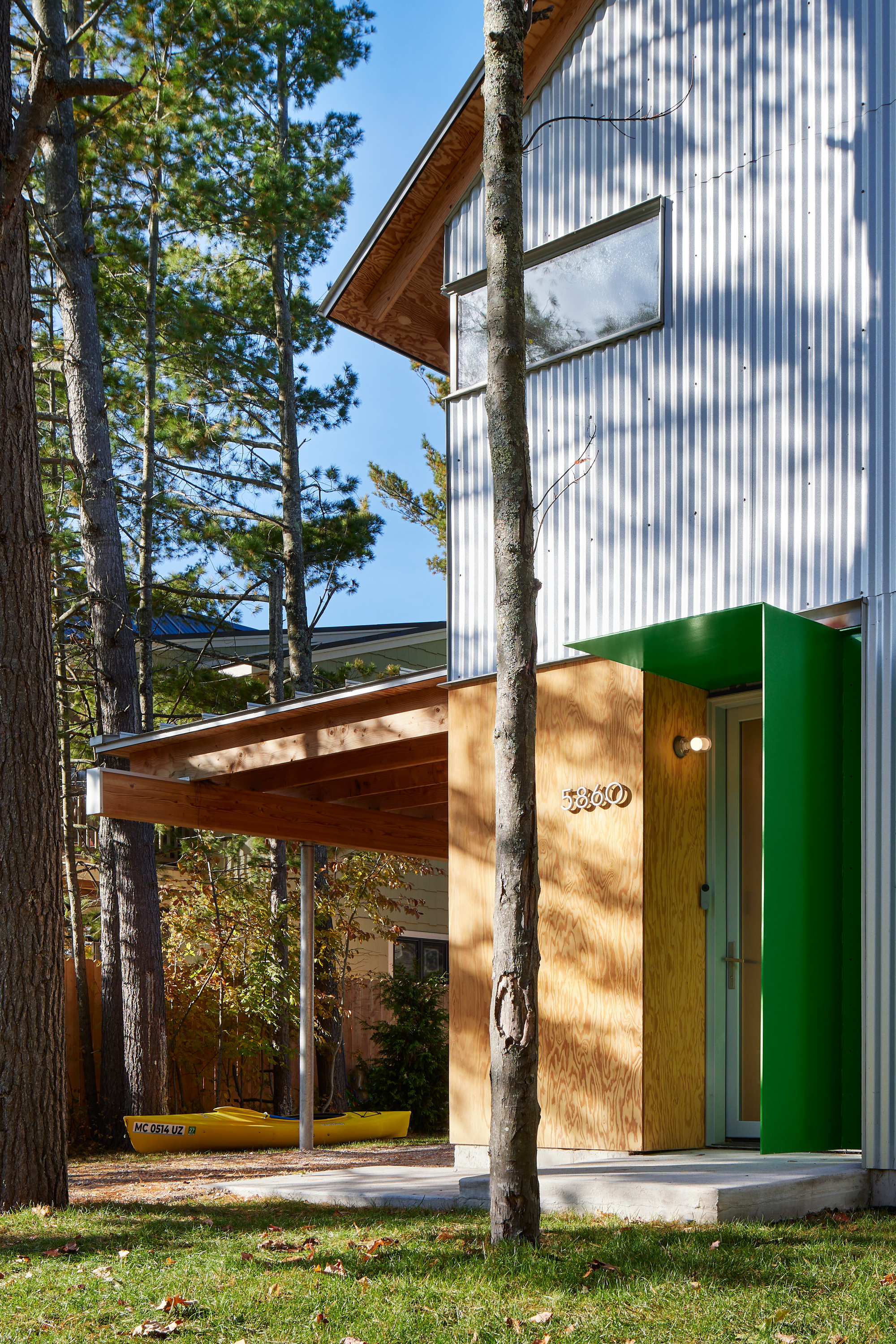 Entry canopy with timber roof and green metal column among pine tree trunks casting shadows