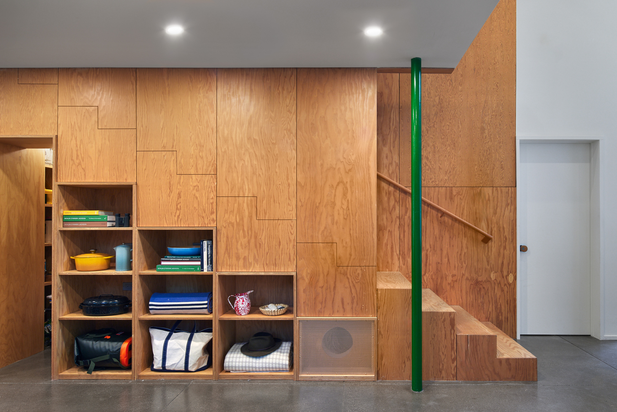 Plywood storage wall with open shelving at base and staircase with green steel handrail