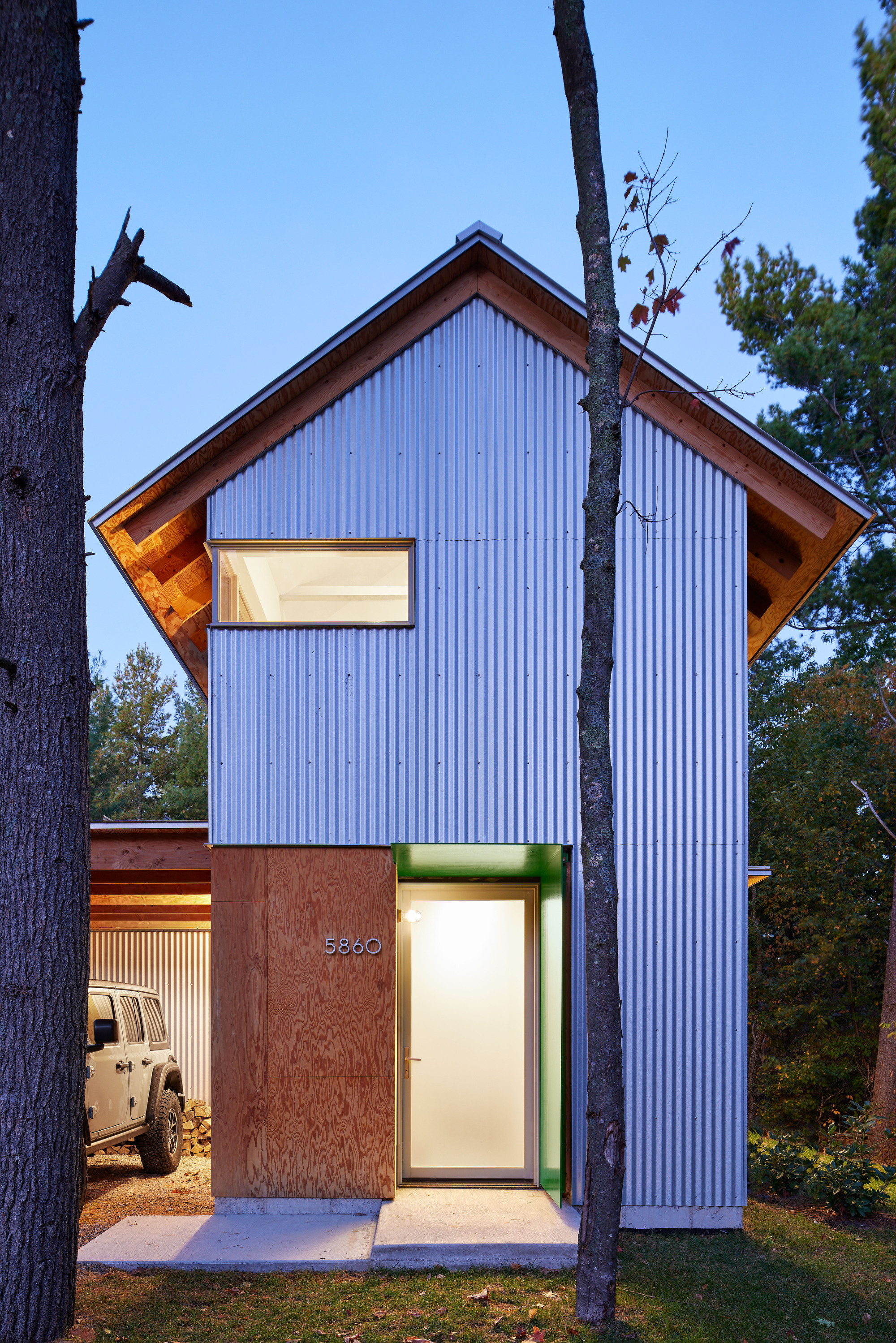Vertical corrugated metal facade with recessed entry and carport framed by pine trees at dusk