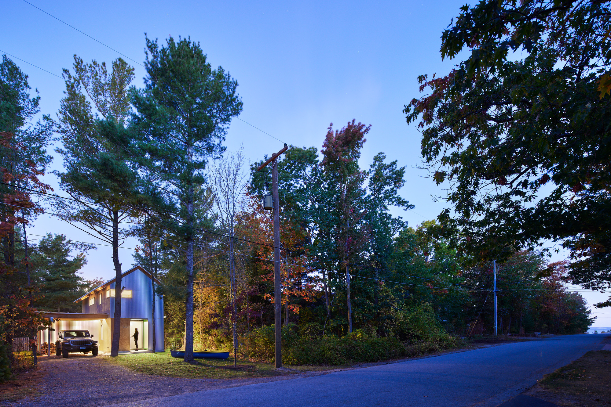 Street view of the gabled volume nestled among trees with illuminated windows at twilight