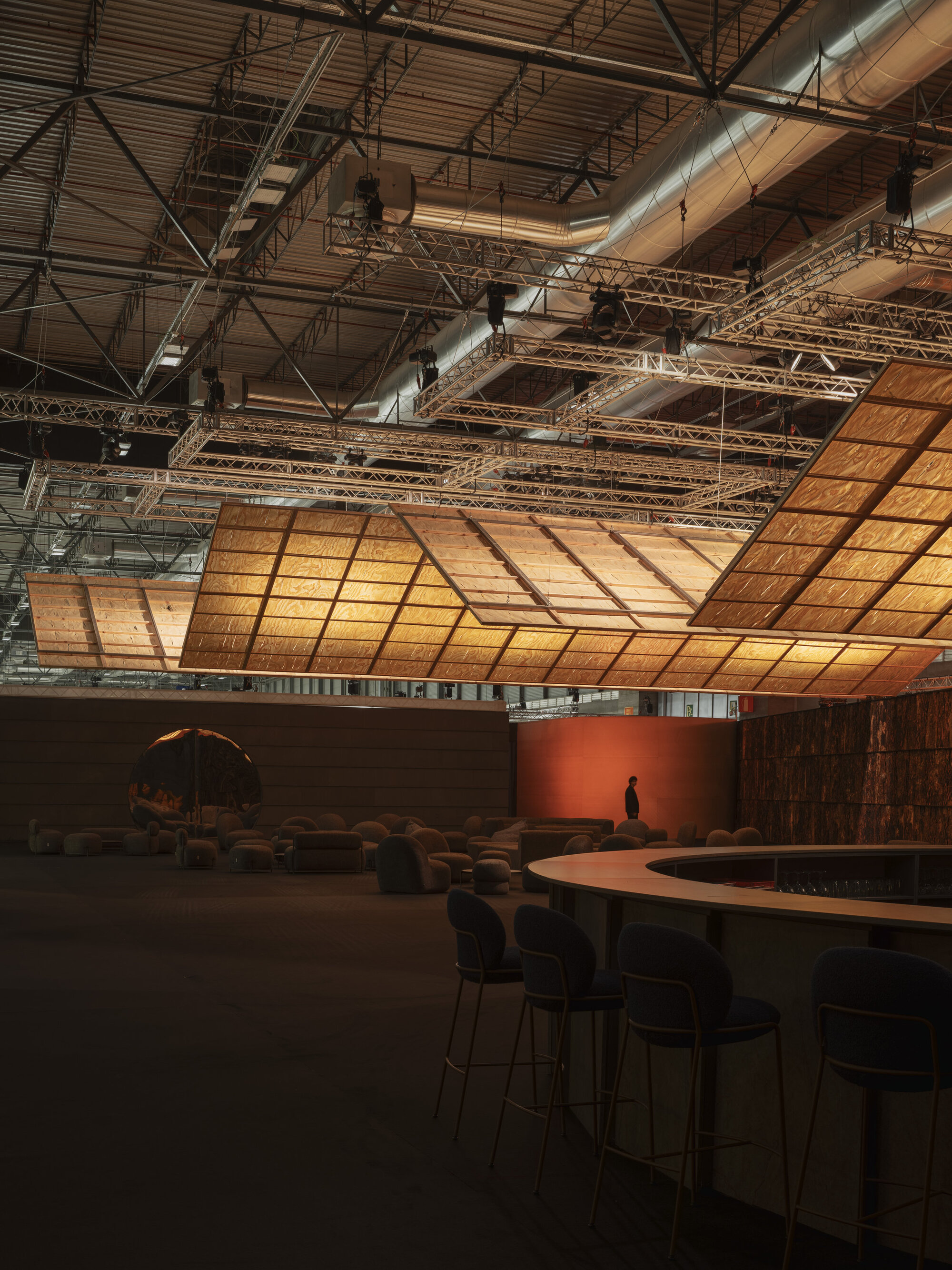 Exhibition hall with curved plywood ceiling, bar counter and a solitary figure near terracotta wall