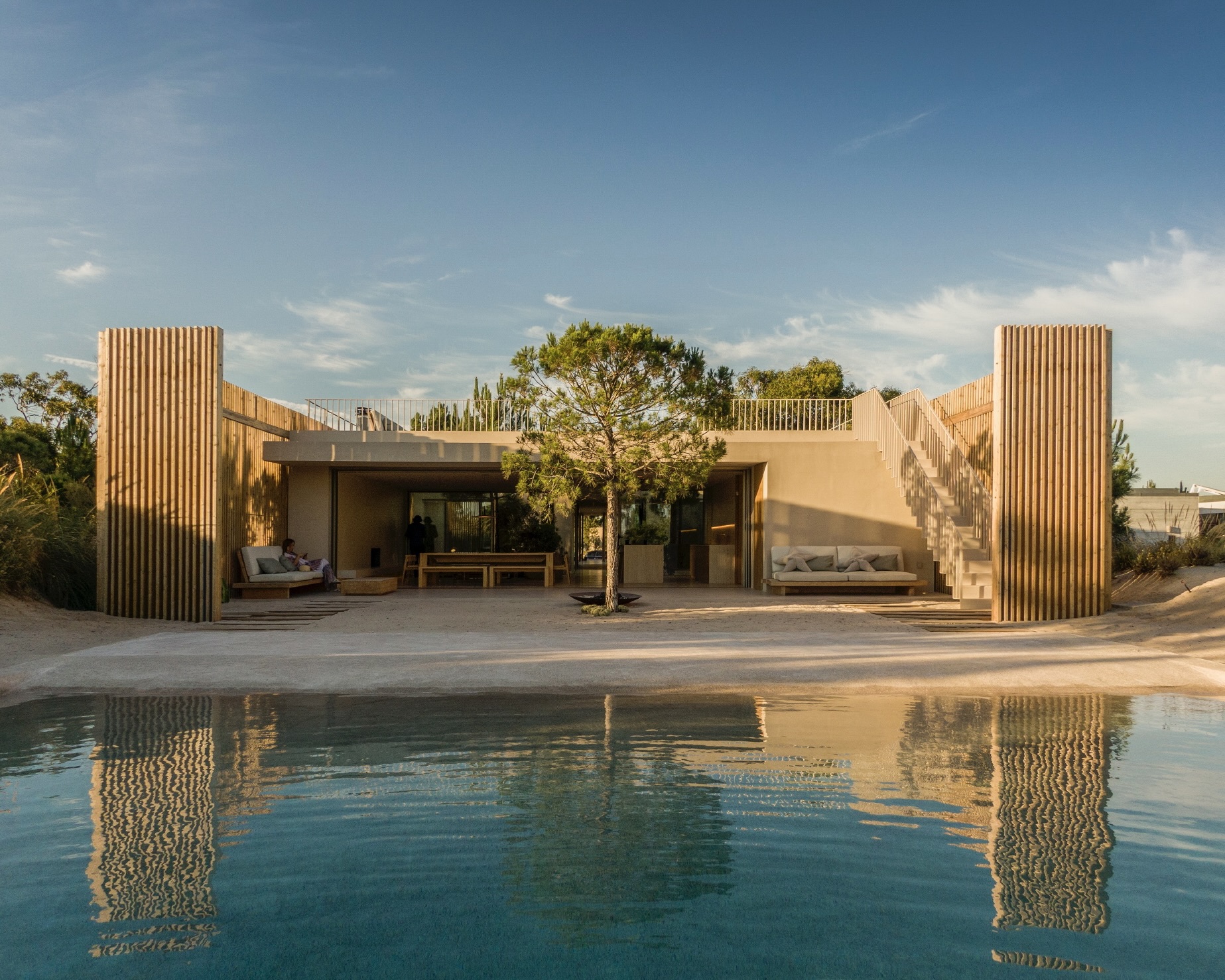 View across the swimming pool toward the timber-slatted facade and terrace in afternoon light