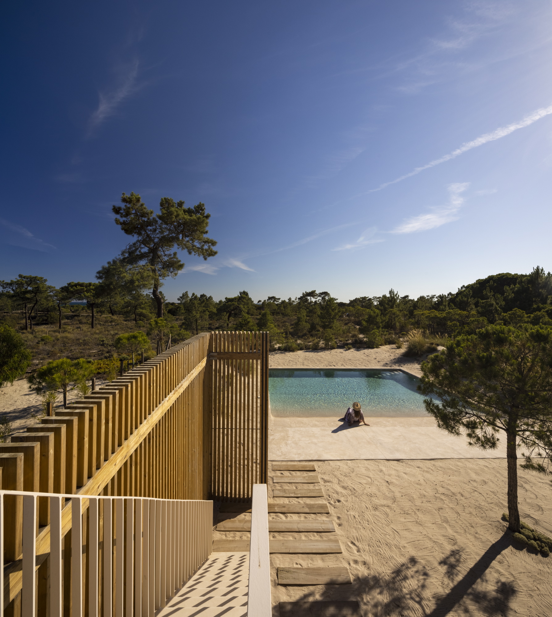 Timber slatted walkway descending toward a pool and sandy beach with pine trees under clear blue sky