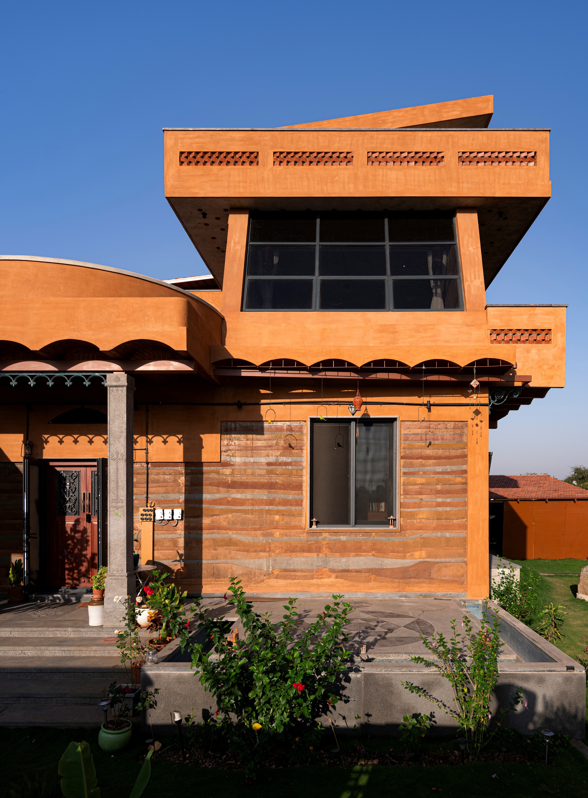 Terracotta brick facade with cantilevered upper volume, scalloped overhangs, and potted plants in afternoon light
