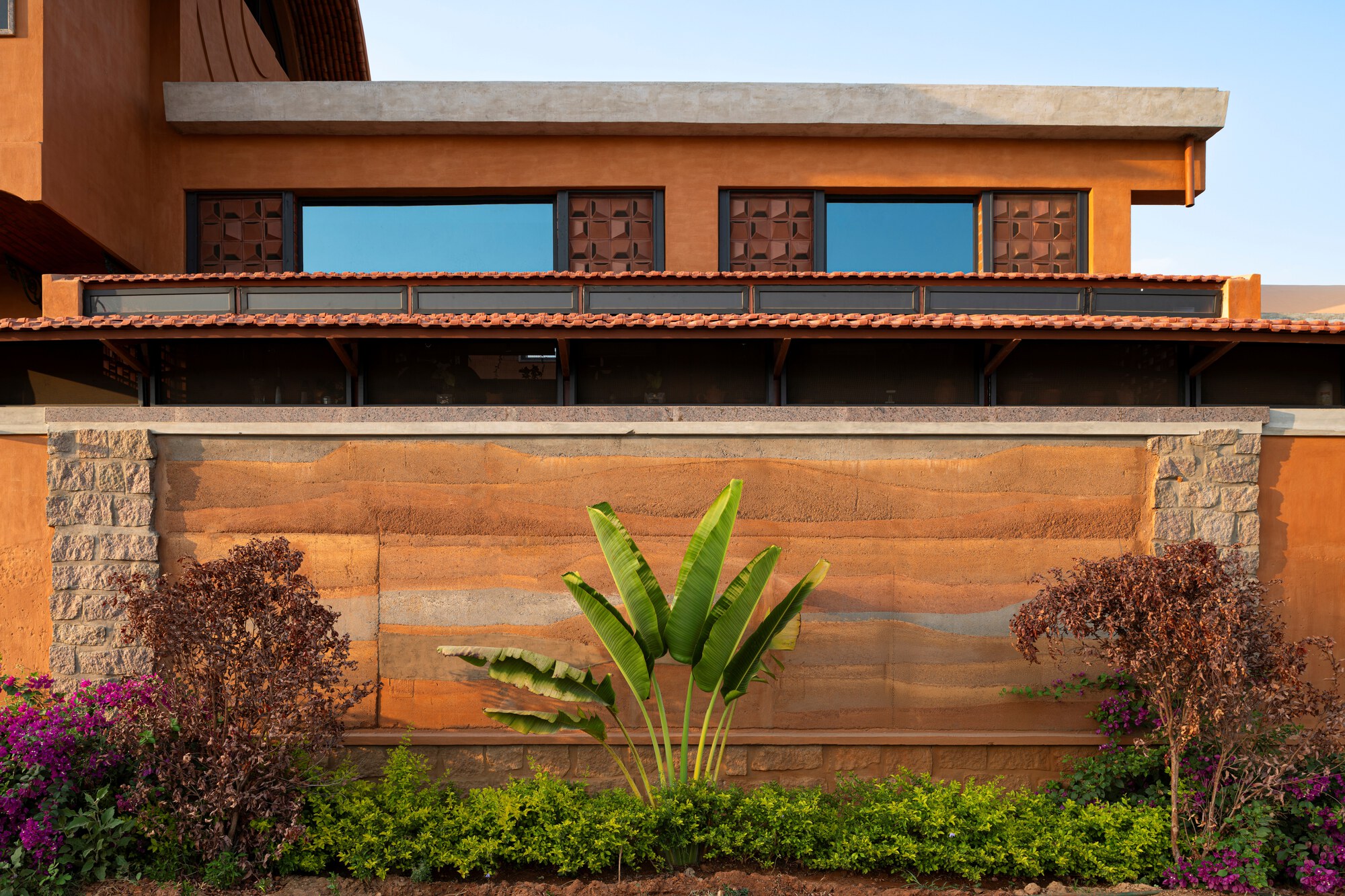 Horizontal banded terracotta wall with stone piers and banana plant against a hedge