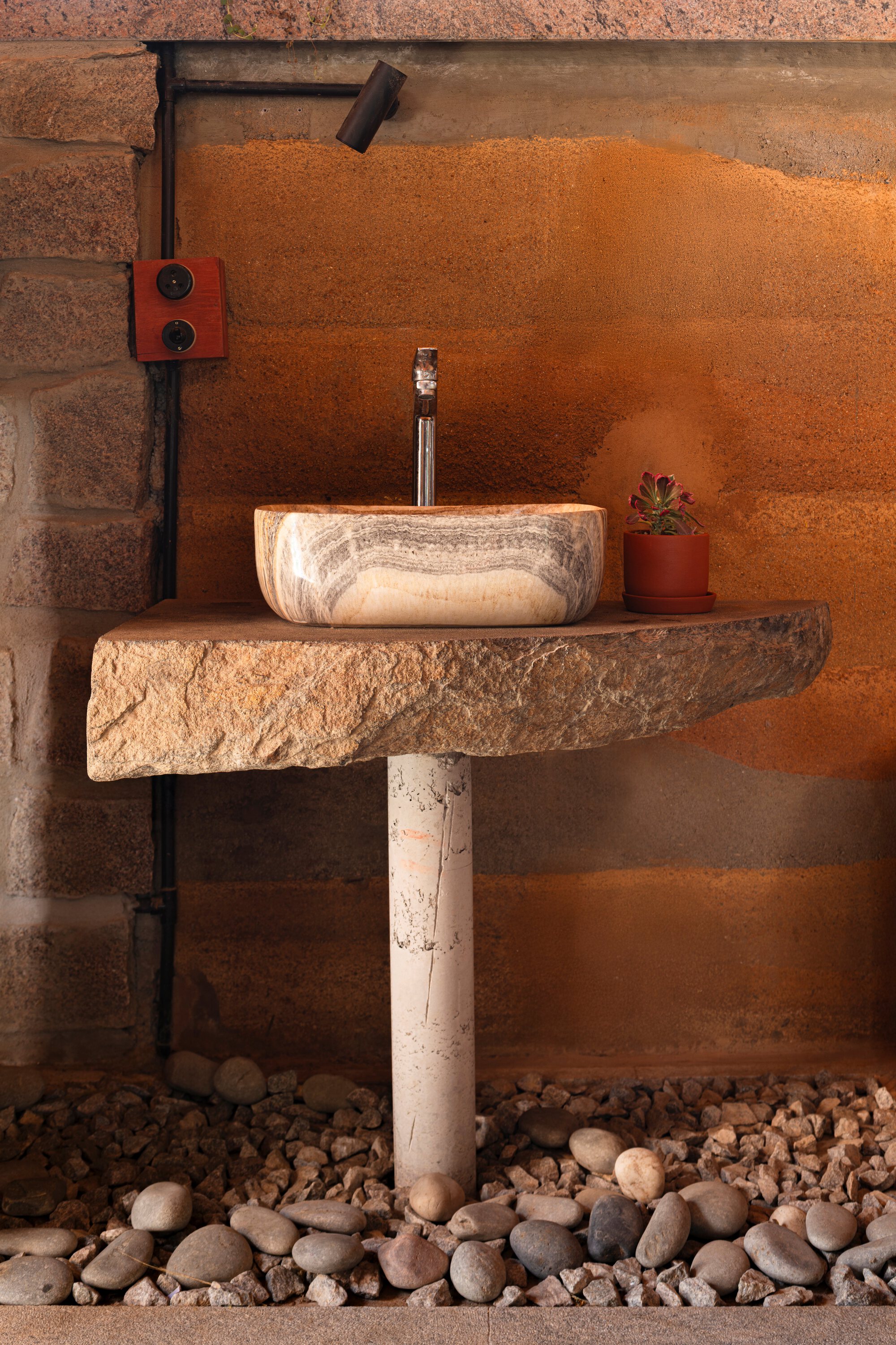 Stone vessel sink on rough-edged counter above river pebble floor with exposed plumbing and terracotta wall