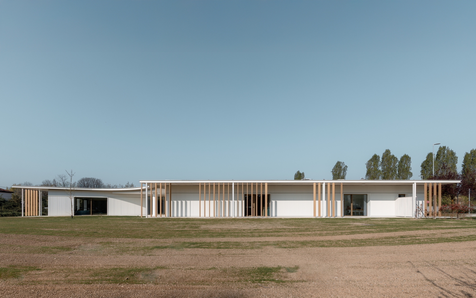Long facade showing alternating white panels and timber screens facing an open field with distant trees