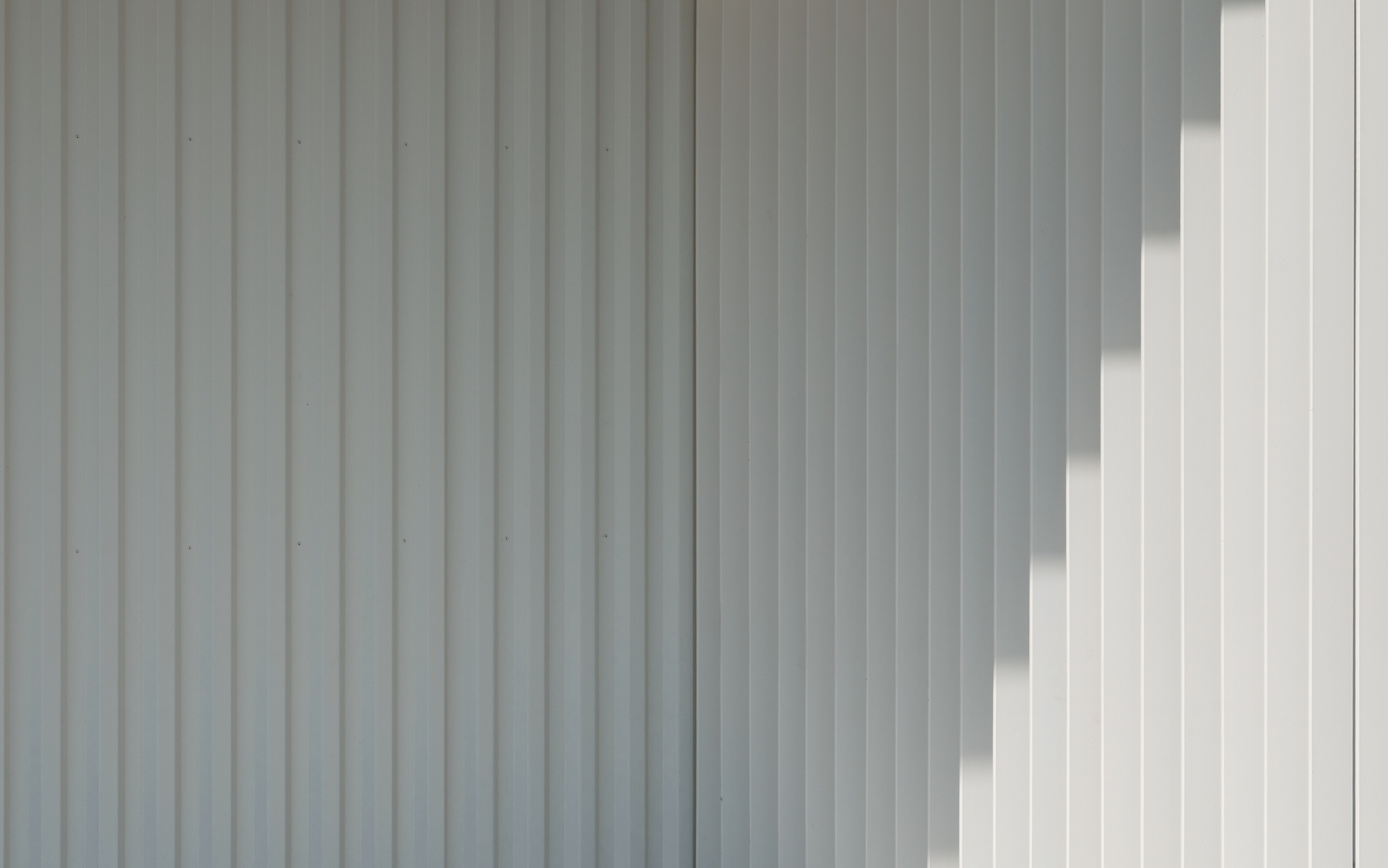 Close-up of vertical corrugated metal cladding in light grey with diagonal sunlight shadows across the surface