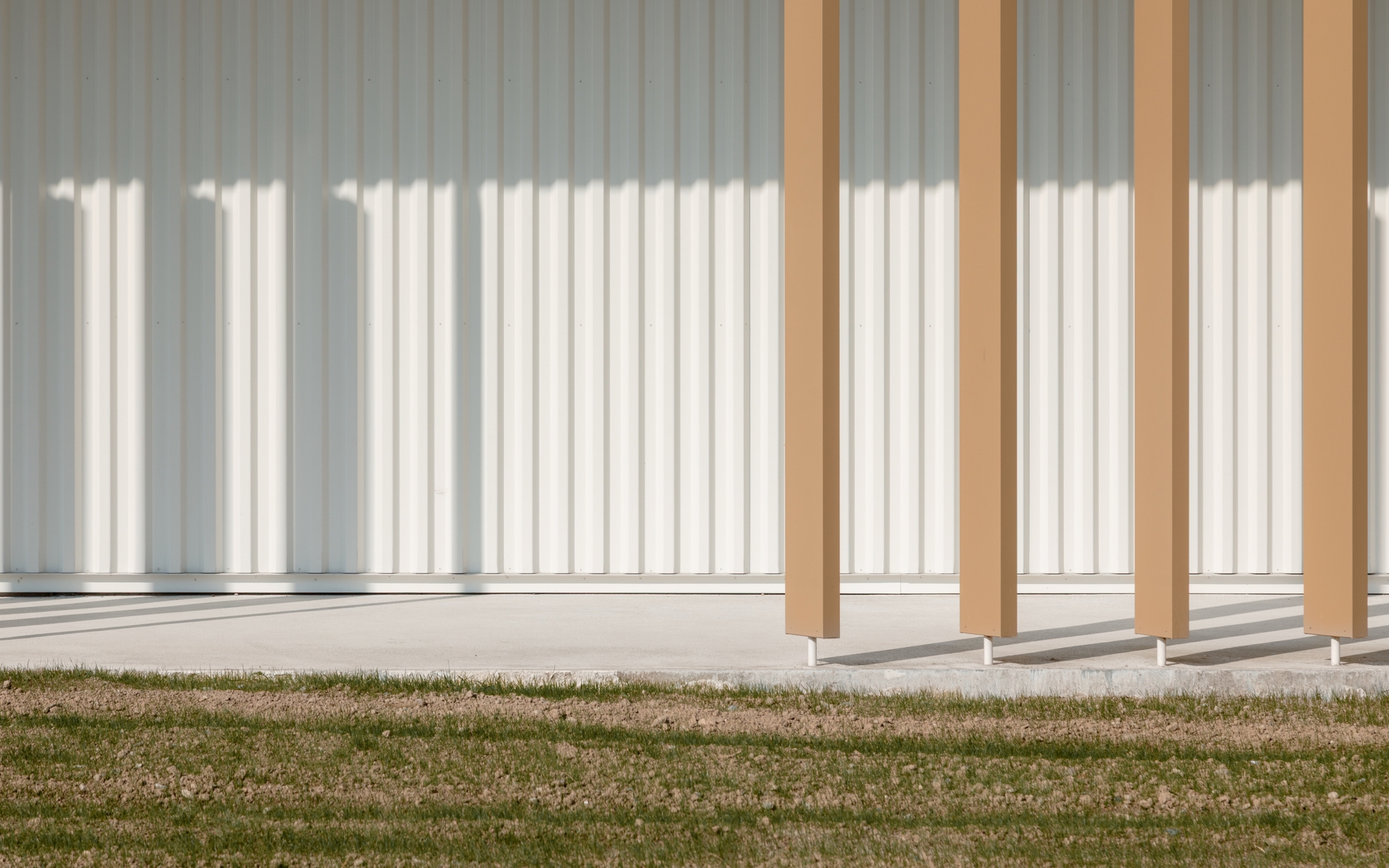 Timber colonnade with square columns casting sharp shadows onto lawn and corrugated metal facade behind