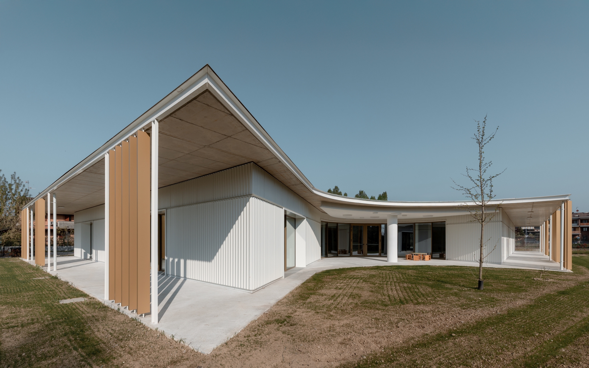 Exterior view showing the gabled and curved roof forms meeting above corrugated walls with a bare tree nearby