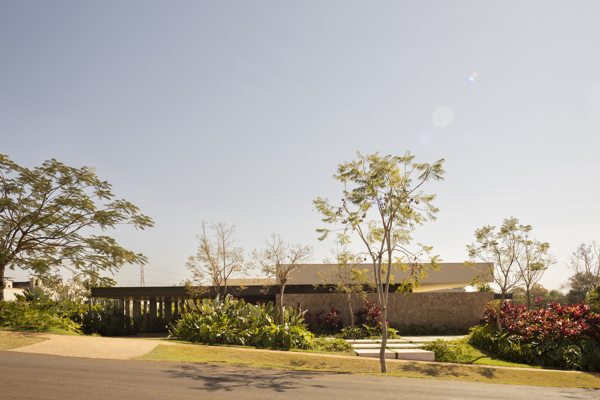 Low horizontal volume with timber colonnade framed by young trees and tropical plantings along a curved driveway