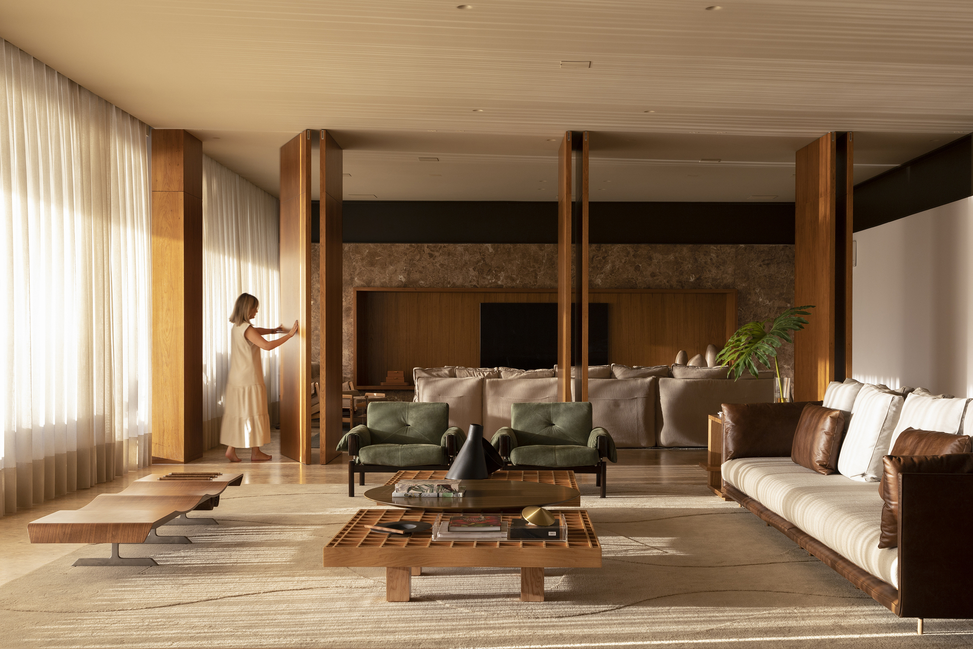 Living room with timber columns and slatted ceiling as a figure adjusts sheer curtains near the glazed wall