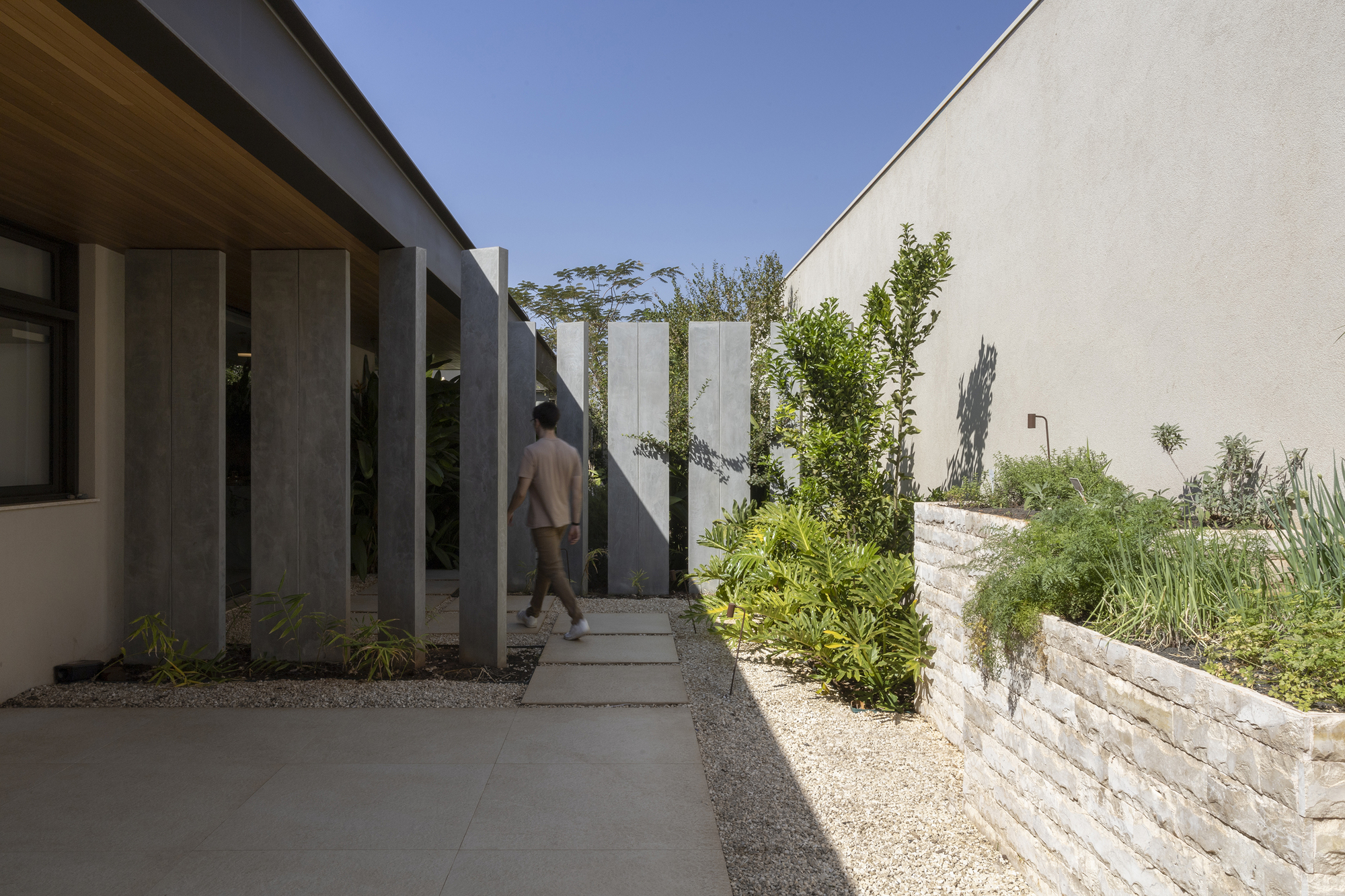 Exterior walkway with vertical metal fins alongside a white stone retaining wall and figure walking