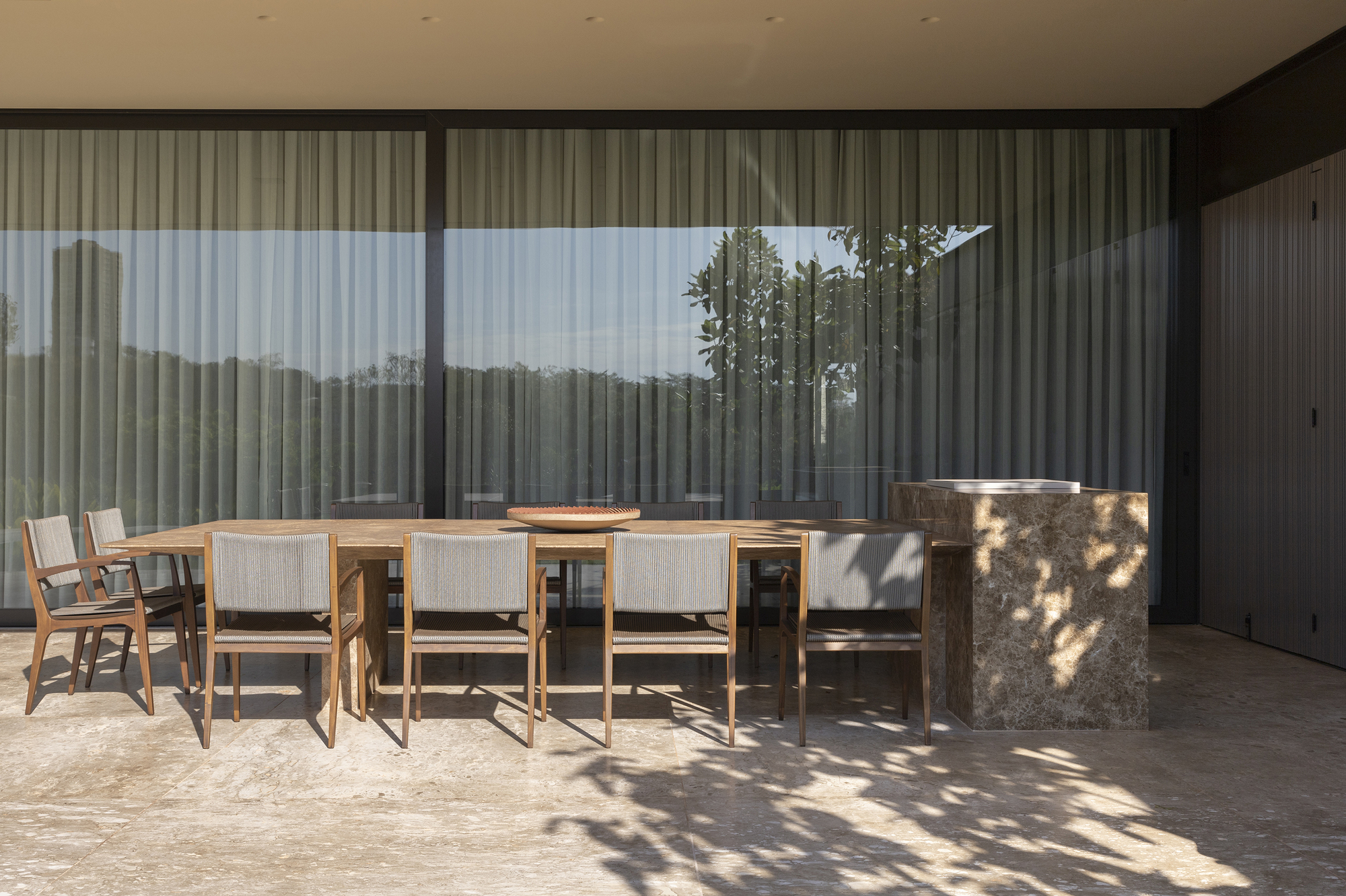 Covered terrace with dining table and chairs casting dappled shadows under tree canopy beyond glass
