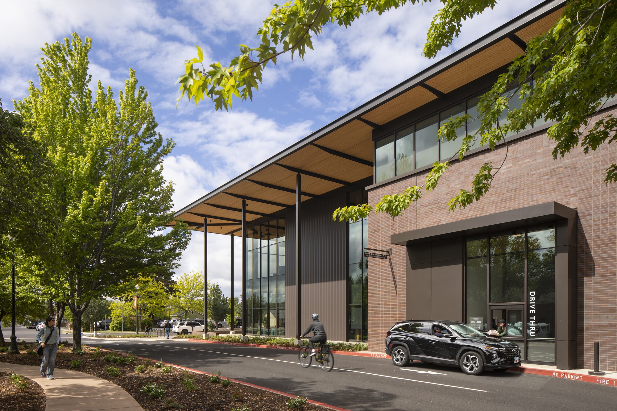 Corner view showing brick masonry volumes and steel canopy framing beneath leafy tree branches